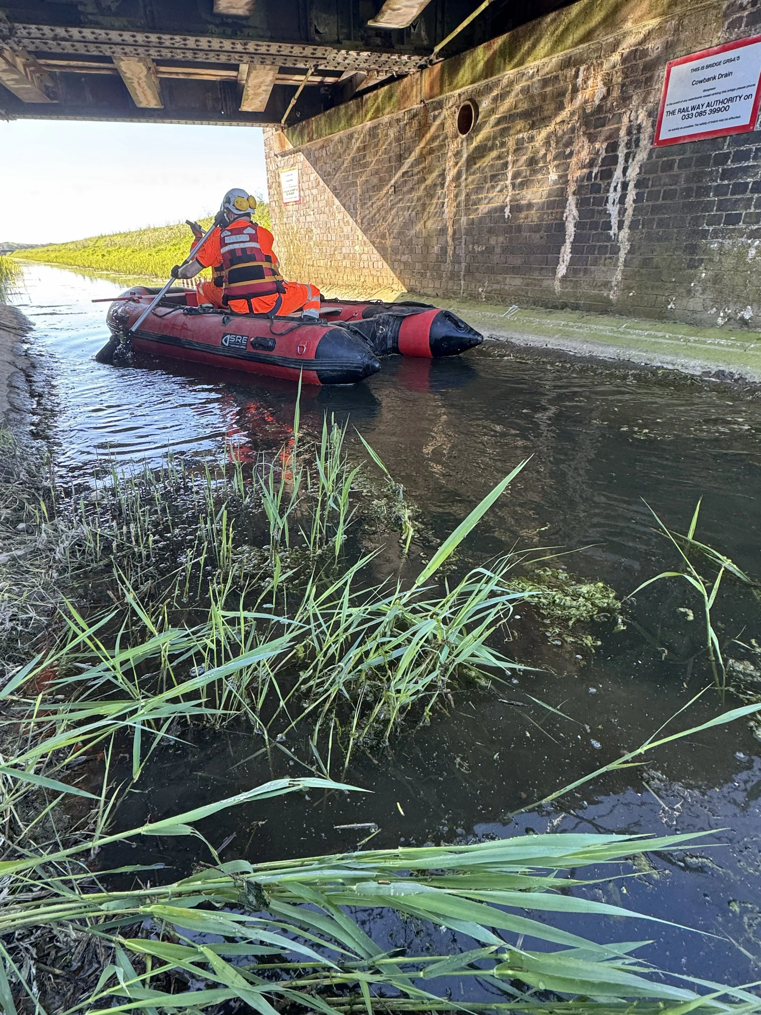 A person in an orange safety suit paddles a small red inflatable boat through a waterway under a bridge. The person wears a helmet with a light and is looking at a phone. The brick wall of the bridge has signs about the Cowdrain Drain and railway aut