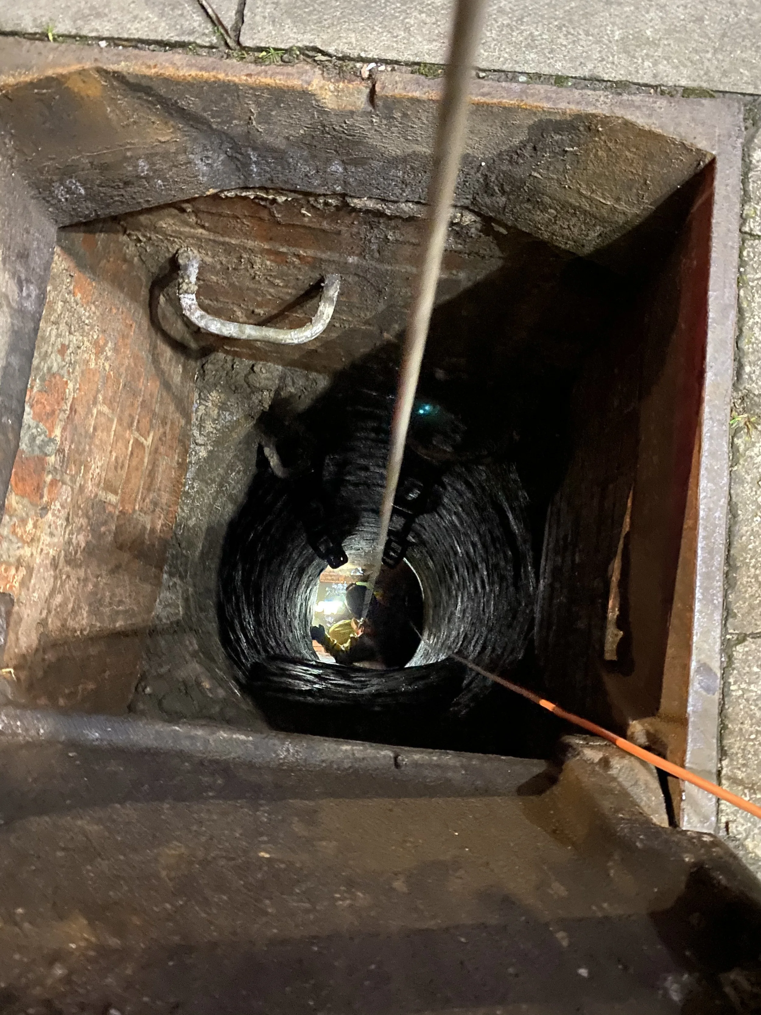 Looking down into a narrow, brick-lined underground inspection or utility hole with a ladder. Two people are visible at the bottom, working or inspecting, with tools and a flashlight.