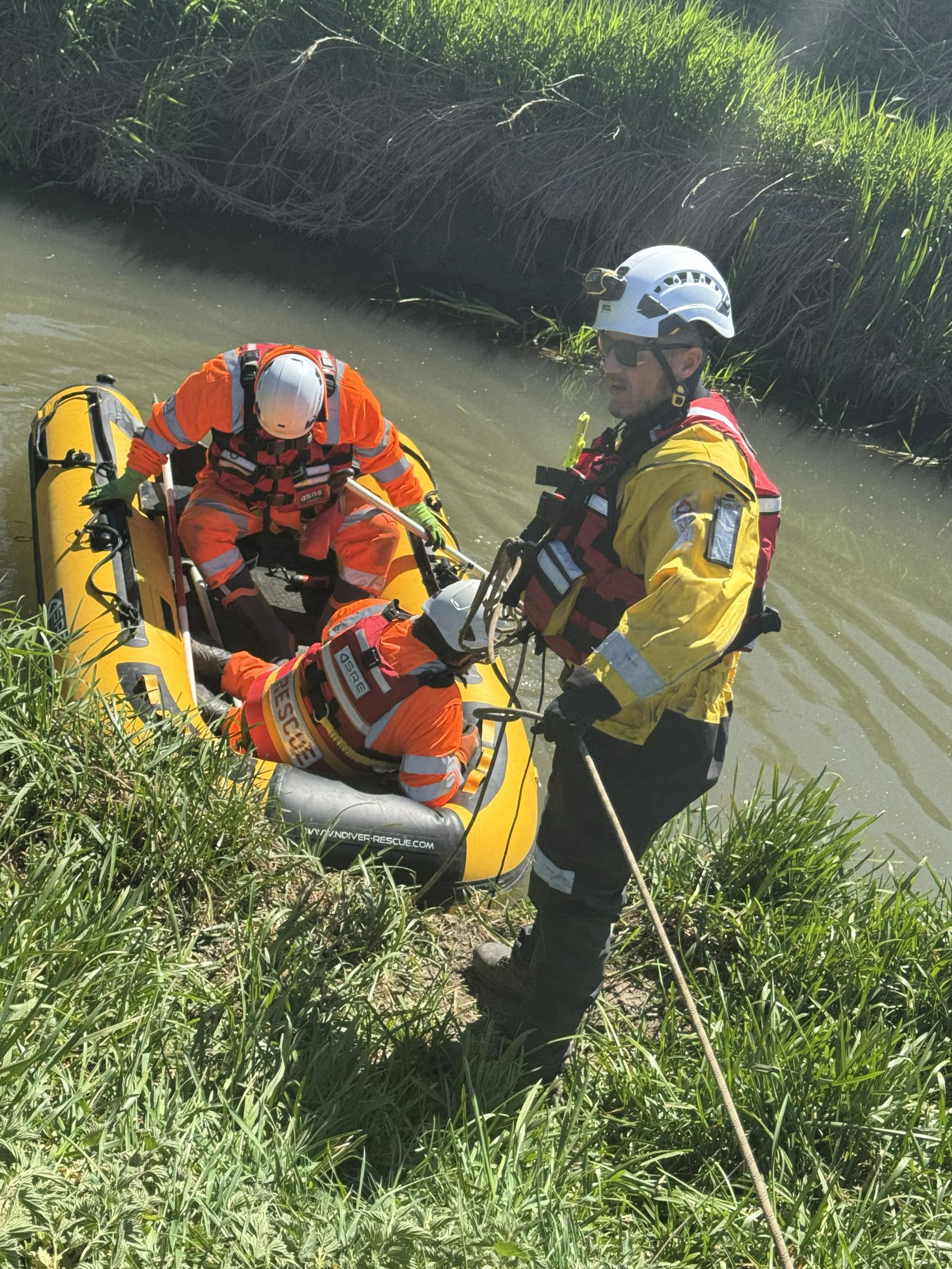 Rescue workers in orange and yellow uniforms assist a person from a yellow inflatable boat near a grassy riverbank.