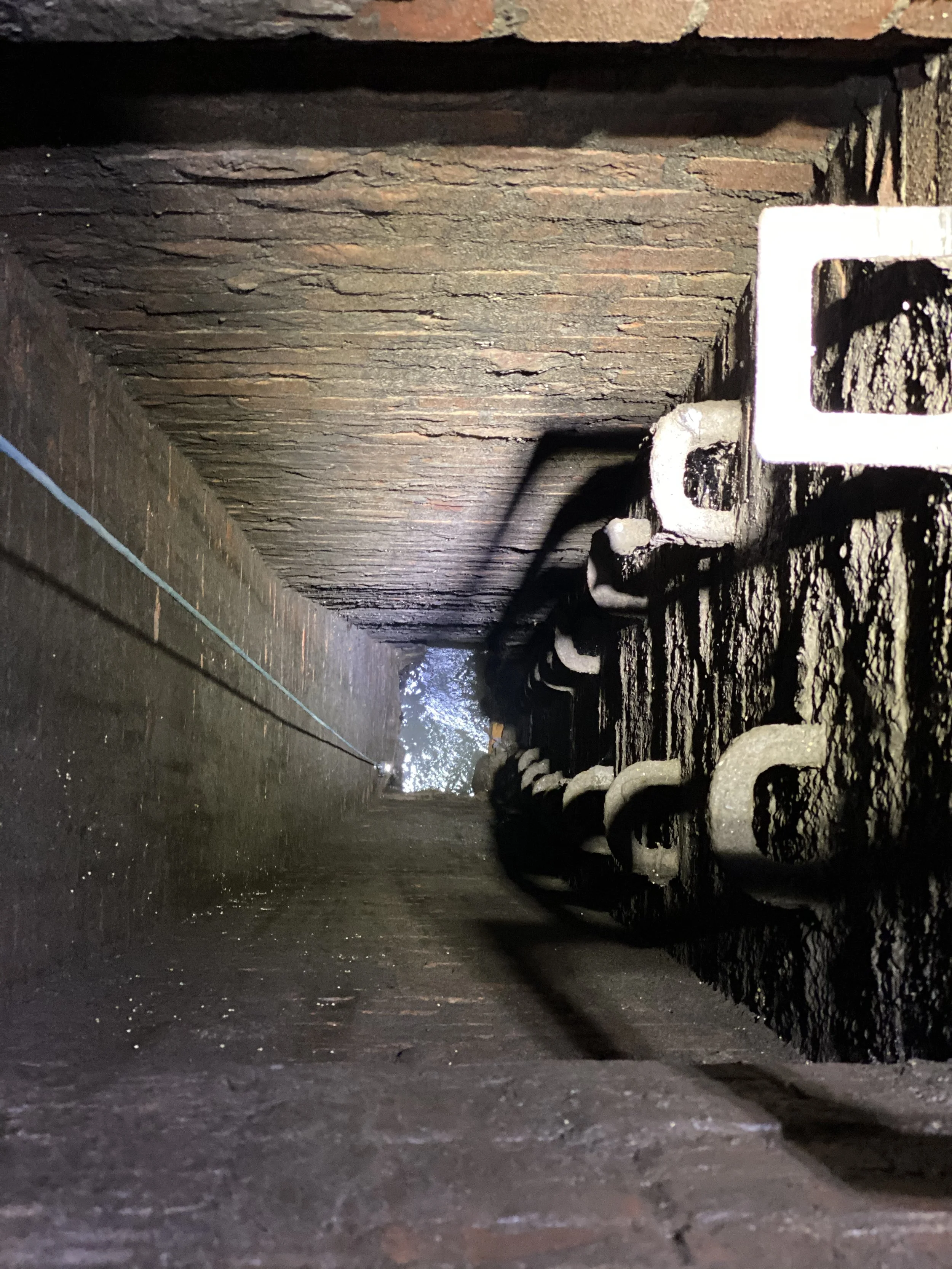 Looking through a narrow brick-lined water tunnel with pipes and a small stream of flowing water.