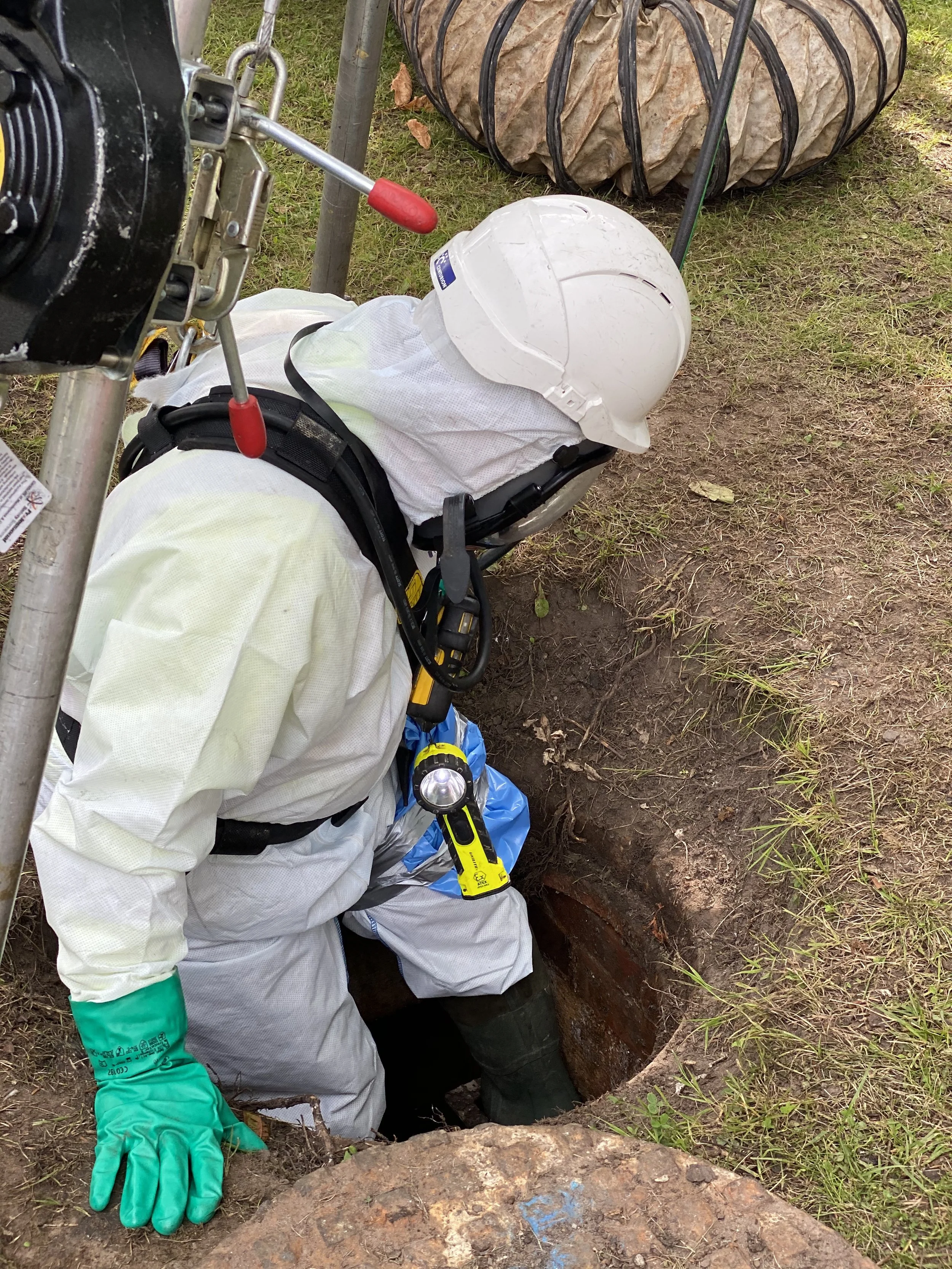 Worker wearing protective gear, including a white suit, helmet, and gloves, inspecting or entering a manhole during a sewer or infrastructure maintenance job.