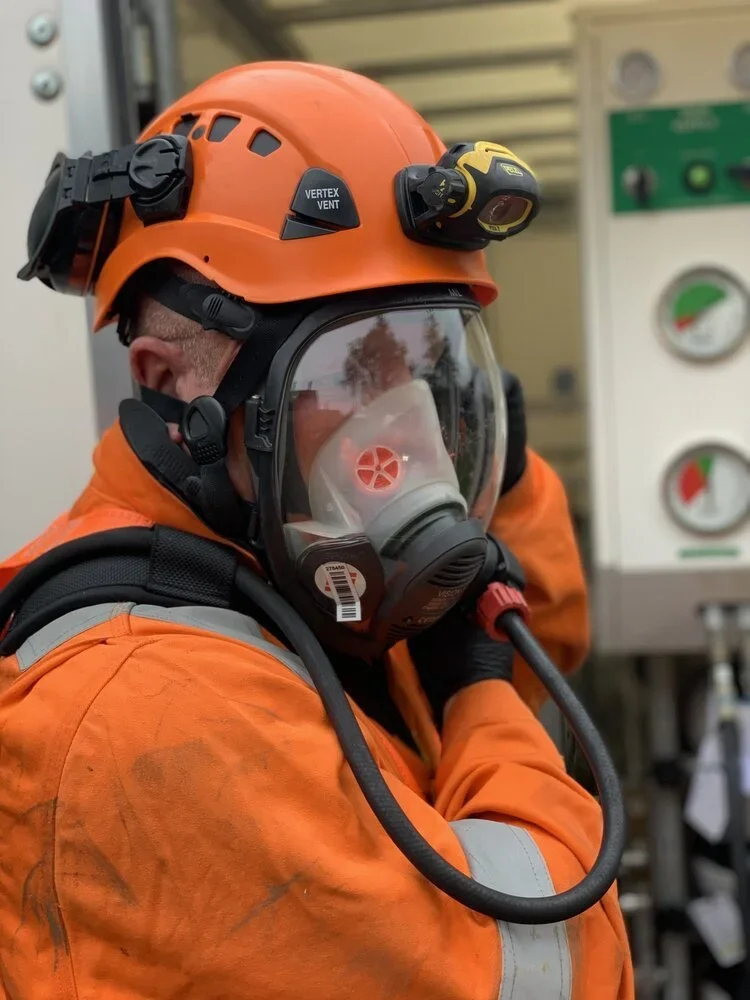 Close-up of an industrial worker wearing an orange safety helmet with headlamp, a clear full-face respirator, and orange protective jacket in a technical environment.