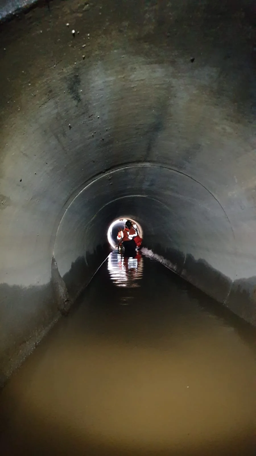 Construction workers in safety vests inspecting a flooded tunnel with water on the floor and light at the far end.