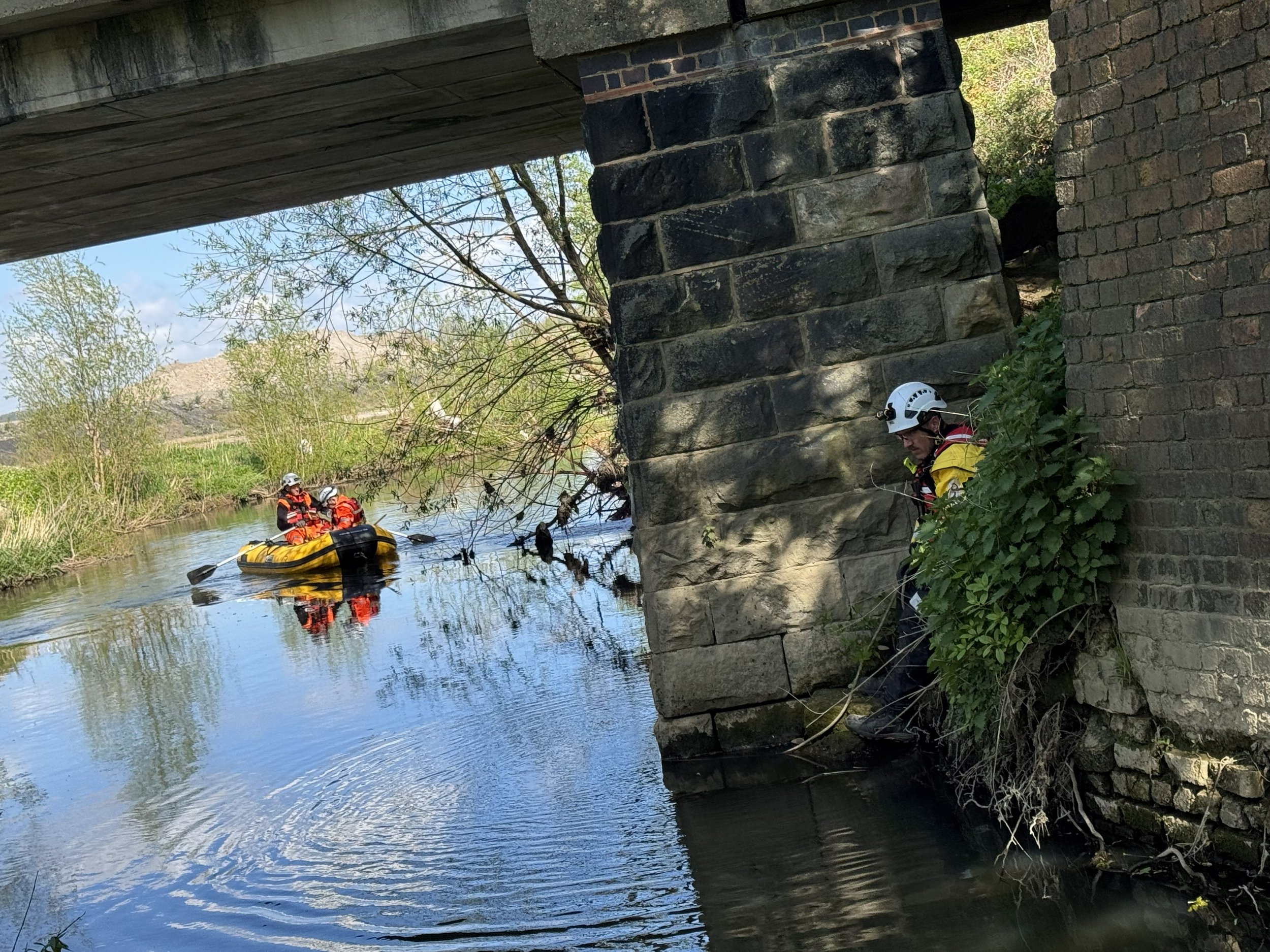 Two rescue workers in yellow and orange life jackets and safety helmets are in a small boat on a calm river, under a bridge with large stone and brick supports, surrounded by green trees and bushes.
