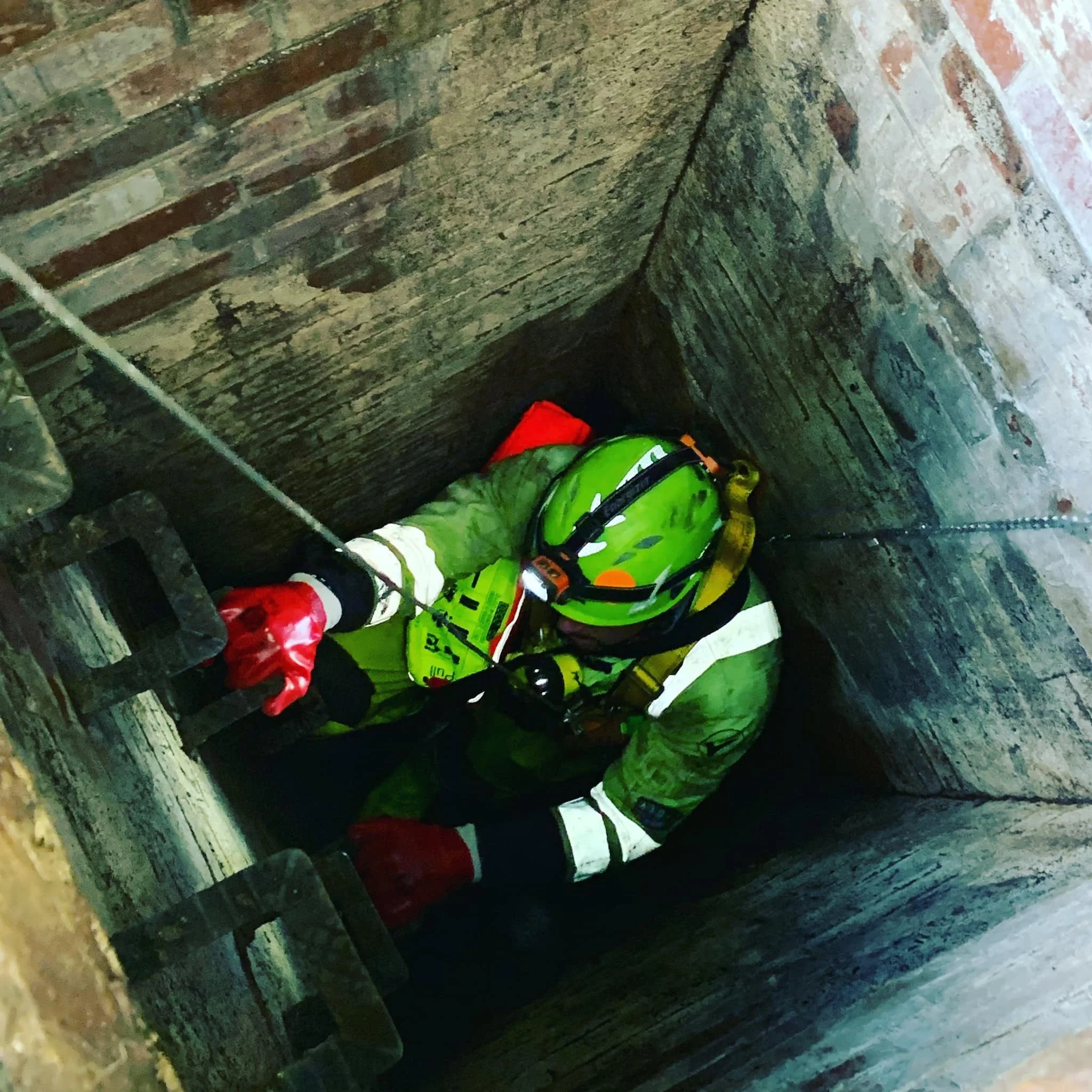 A firefighter wearing a bright green helmet, reflective protective clothing, and red gloves is inside a narrow vertical confined space, possibly a utility or maintenance shaft, holding onto a safety rope.
