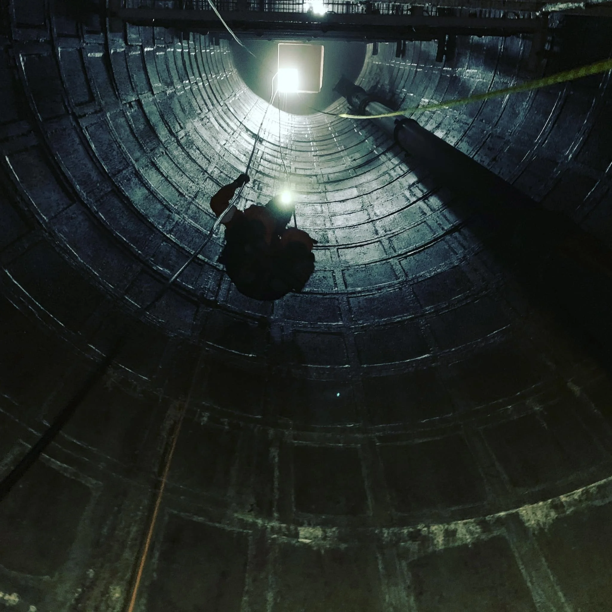 A person working inside a large, cylindrical industrial tank, possibly cleaning or inspecting, with the tank opening visible at the top and light coming from above.