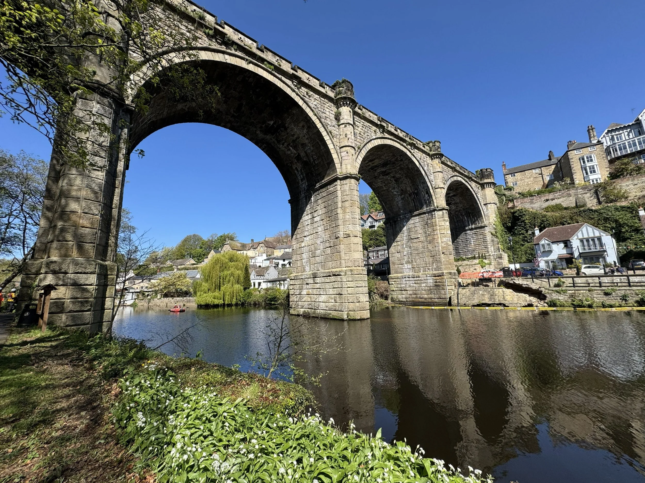 Stone railway bridge over a river with trees and houses on a hillside in the background under a clear blue sky.