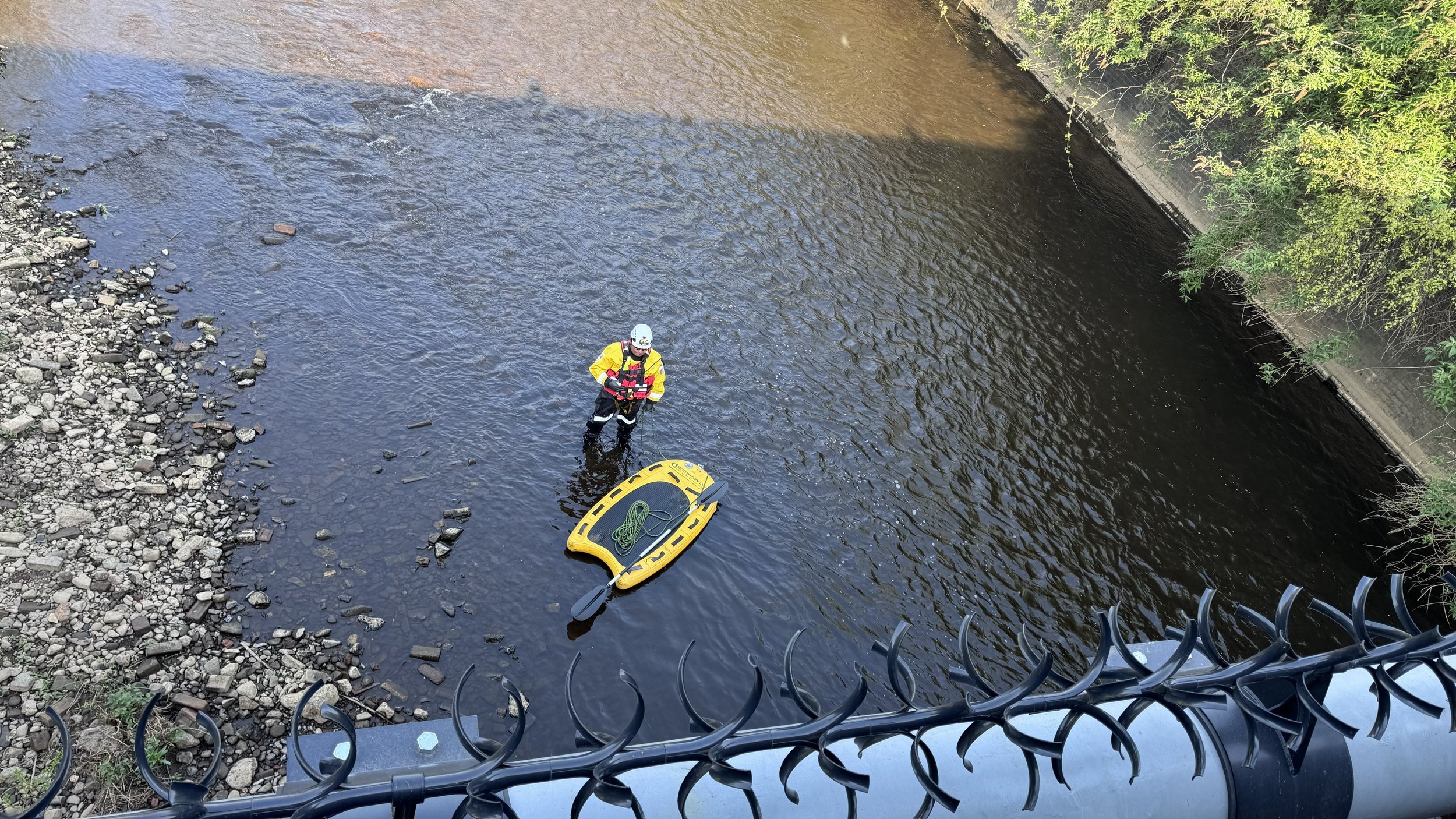 A person wearing a yellow waterproof jacket, black pants, and a white helmet stands in a river near a yellow rescue boat with a green rope. They are holding onto a rope and appear to be involved in a rescue or safety operation. The riverbank is rocky