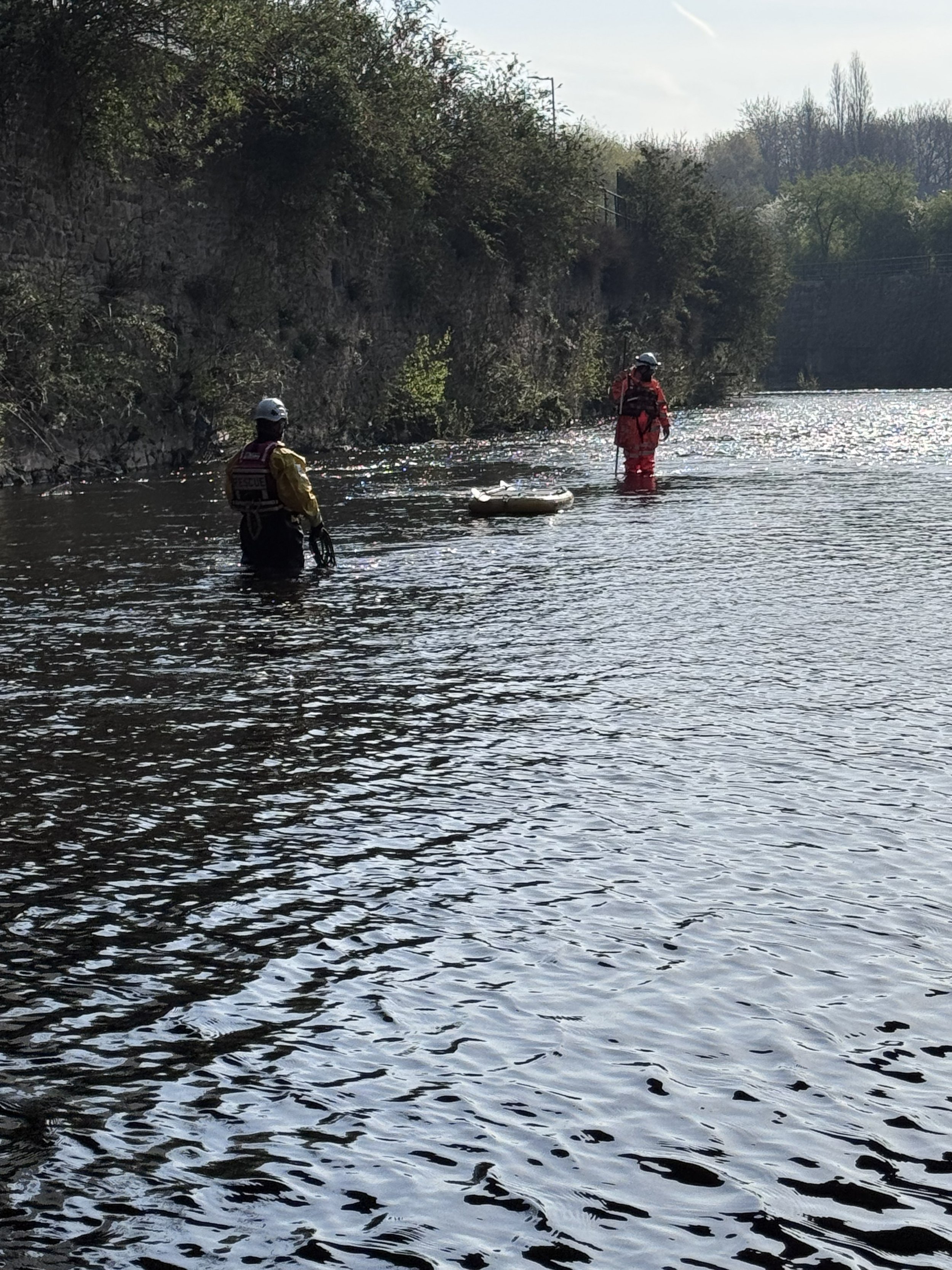 Two rescue workers standing in shallow water near a rocky riverbank, with a small inflatable boat between them, during daytime.