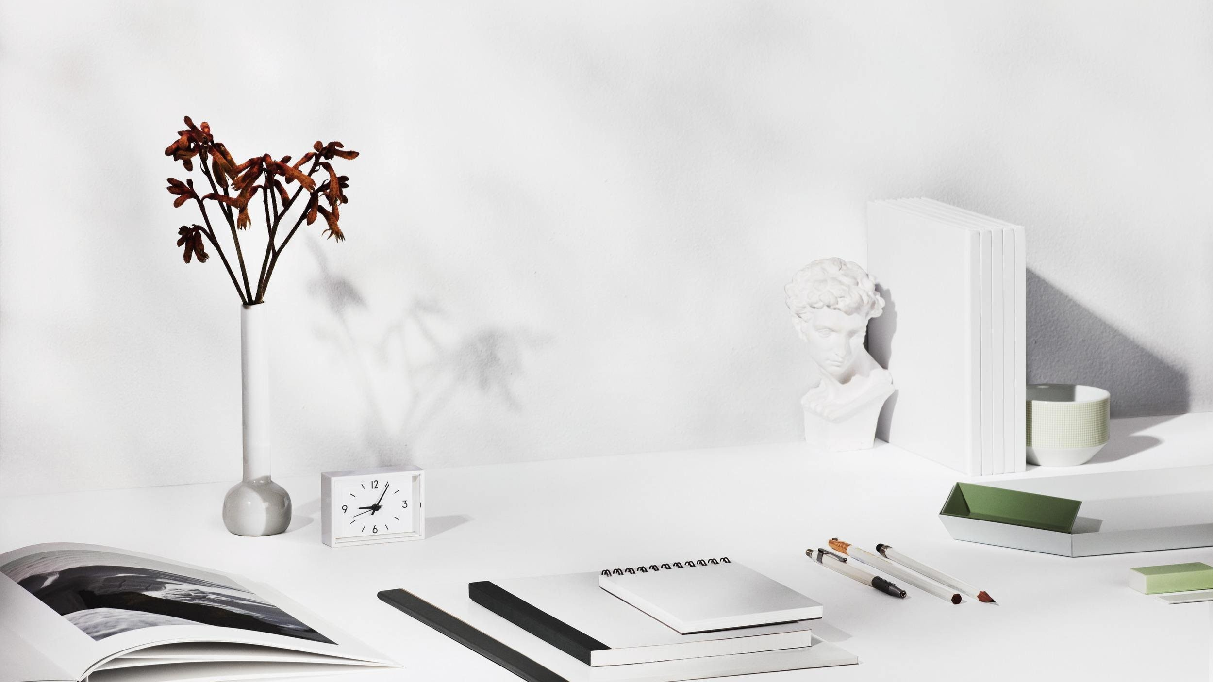 Minimalist white desk with a white vase holding dried flowers, a small white clock, open magazine, black and white notebooks, pens, a white and green organizer, a white sculpture of a woman's bust, a row of white books, a small bowl, and shadowed wall.
