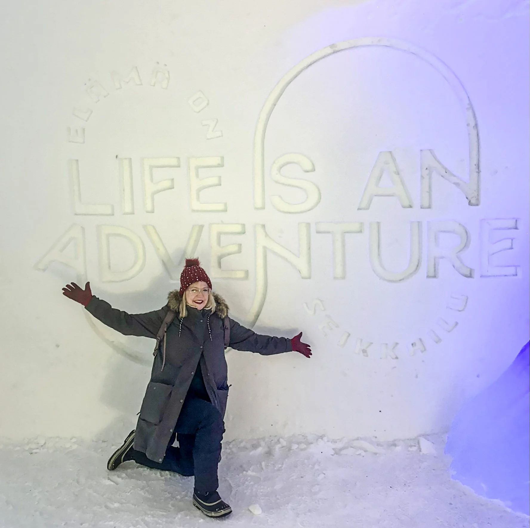 A woman posing in front of a snow wall with the words "LIFE IS AN ADVENTURE" carved into it. She is wearing a dark winter coat, red knit hat with white dots, glasses, gloves, and snow boots, smiling with arms outstretched.