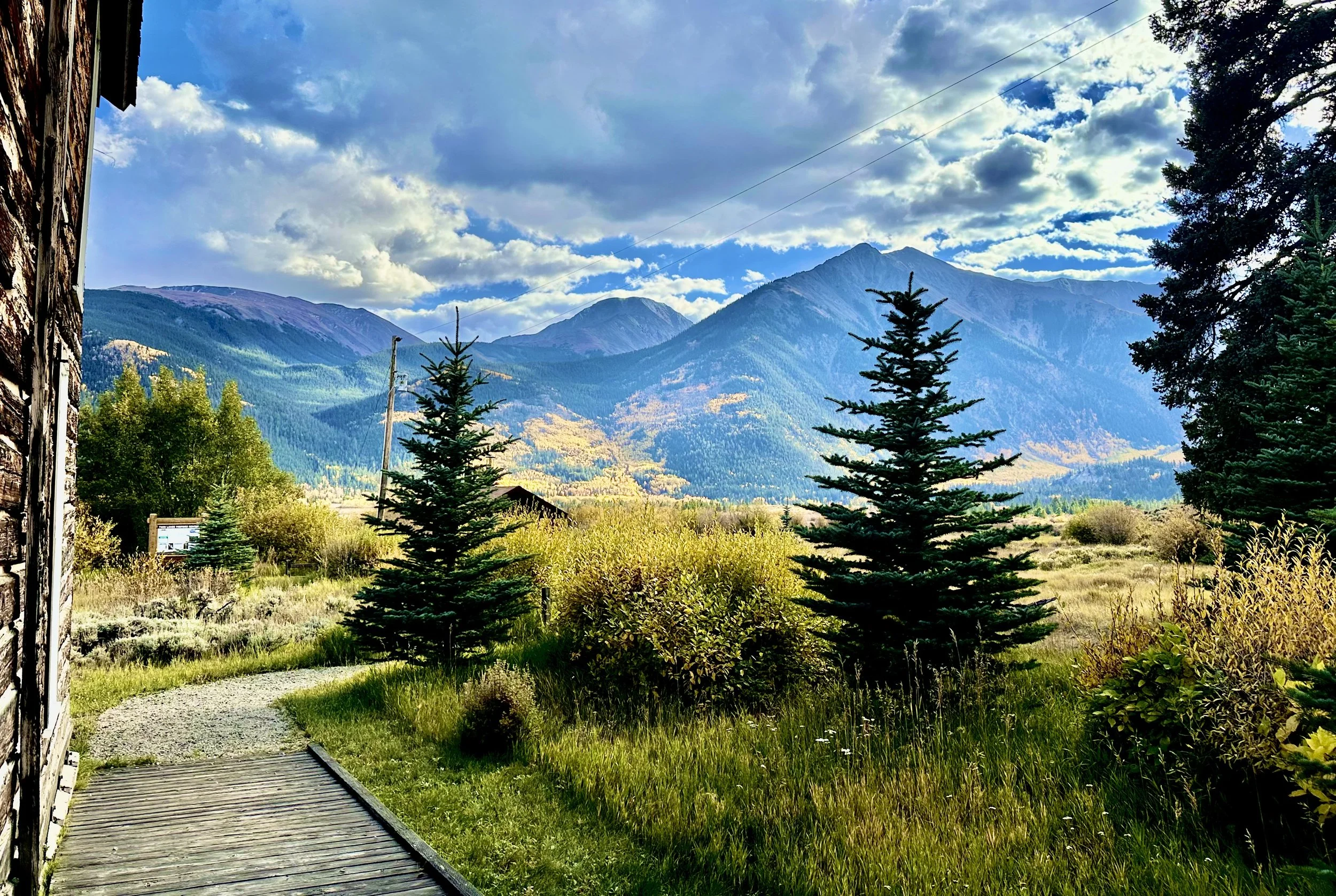 Scenic mountain landscape with green pine trees, grassy area, and a wooden building on the left. Bright sky with clouds and tall mountain peaks in the background.