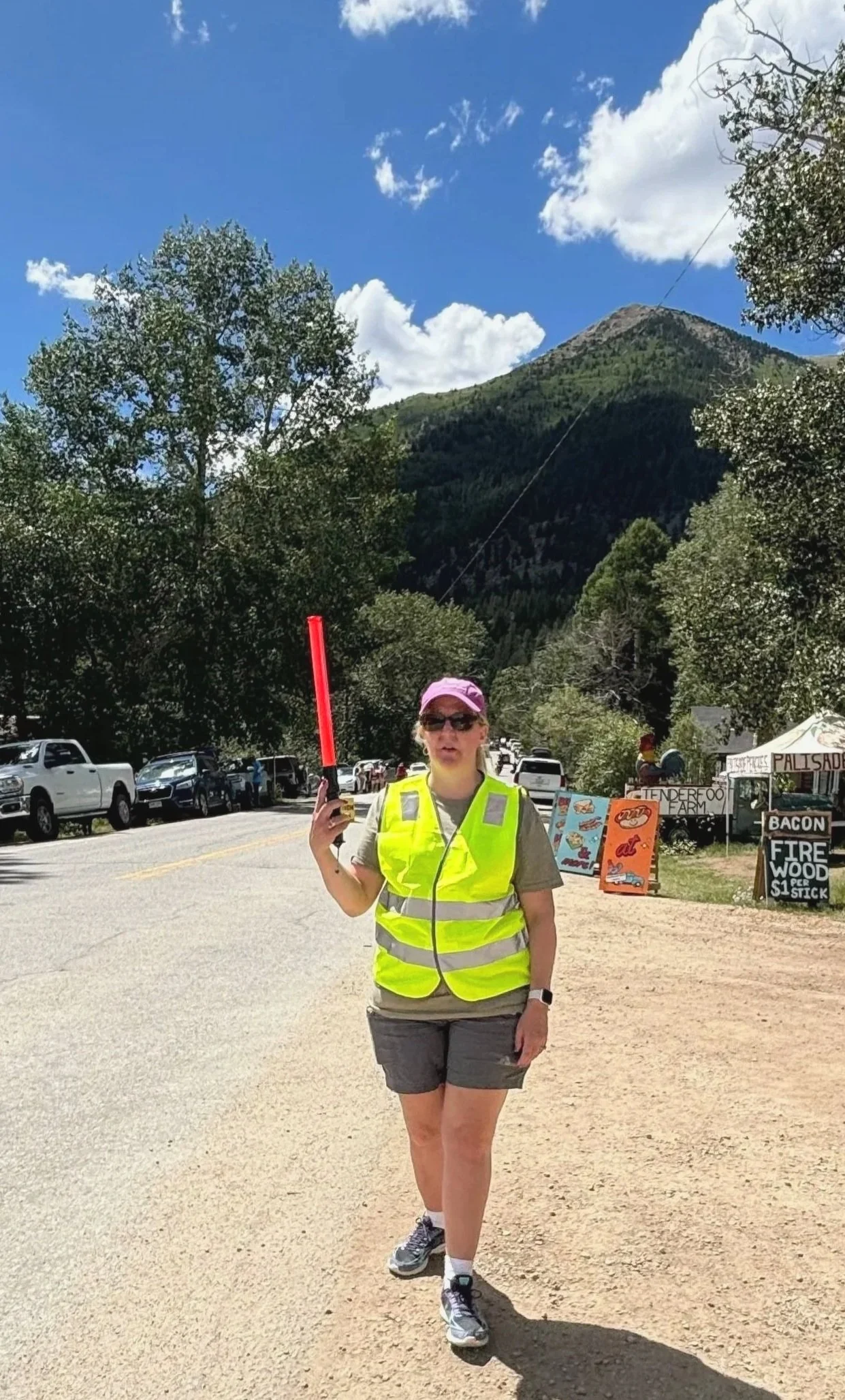 A woman wearing a yellow reflective safety vest, gray shorts, and sneakers, holding a red glow stick standing on a dirt road. Behind her are parked cars, trees, and mountain scenery with blue sky and clouds.