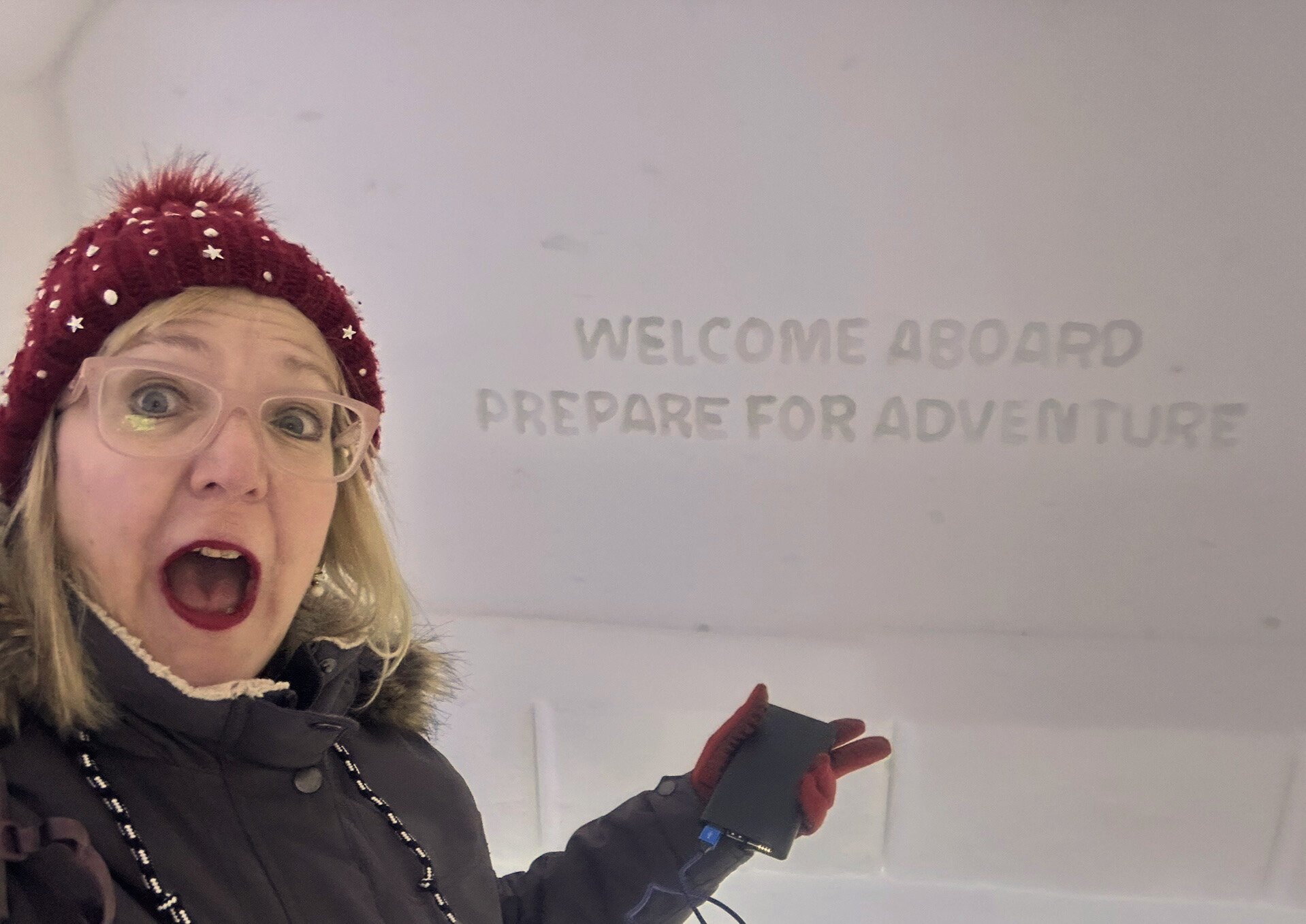 Woman with blonde hair, glasses, red hat with white stars, and black coat holding a device, standing beside a white wall with the message 'Welcome aboard prepare for adventure' written on it.