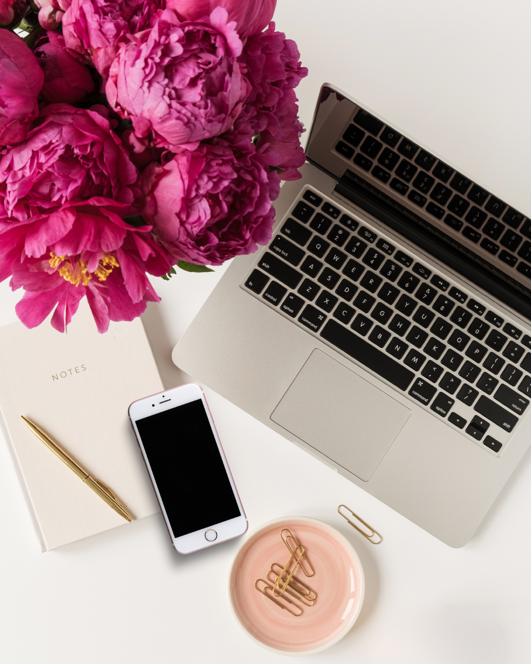 A workspace with a laptop, a pink vase of peonies, a white notebook with a gold pen, an iPhone, a pink bowl with paper clips, and a few loose paper clips.