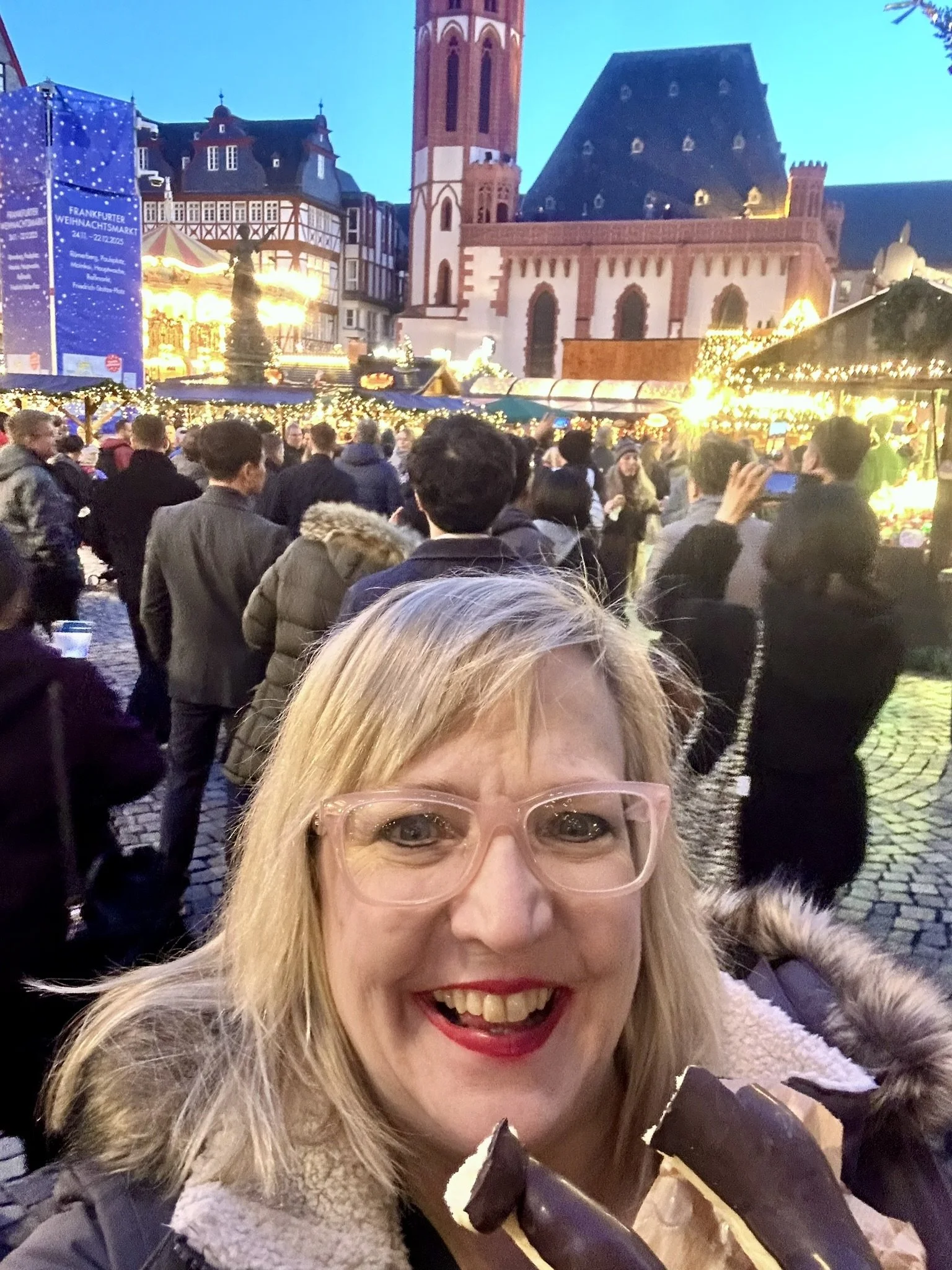 "Woman in pink glasses smiling at camera while holding chocolate at a busy evening Christmas market with glowing lights and crowds in the background