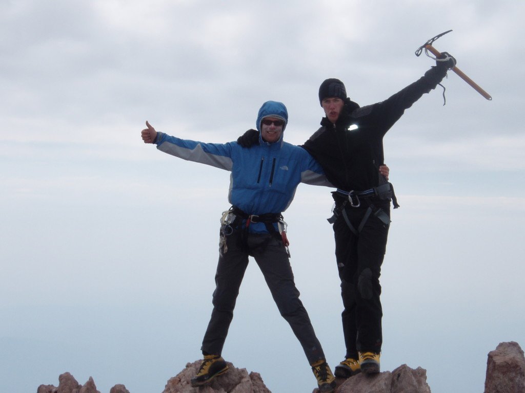 Two climbers standing on rocky summit, celebrating with one giving a thumbs-up and the other raising an ice axe, against a cloudy sky.