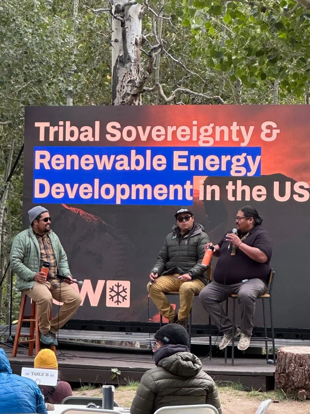 Three men seated on a stage participating in a panel discussion at an outdoor event 