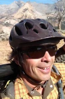 Person wearing a black bicycle helmet and sunglasses, outdoors with mountains in the background.