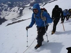 Group of hikers trekking through snowy, mountainous terrain with snow-covered peaks in the background.