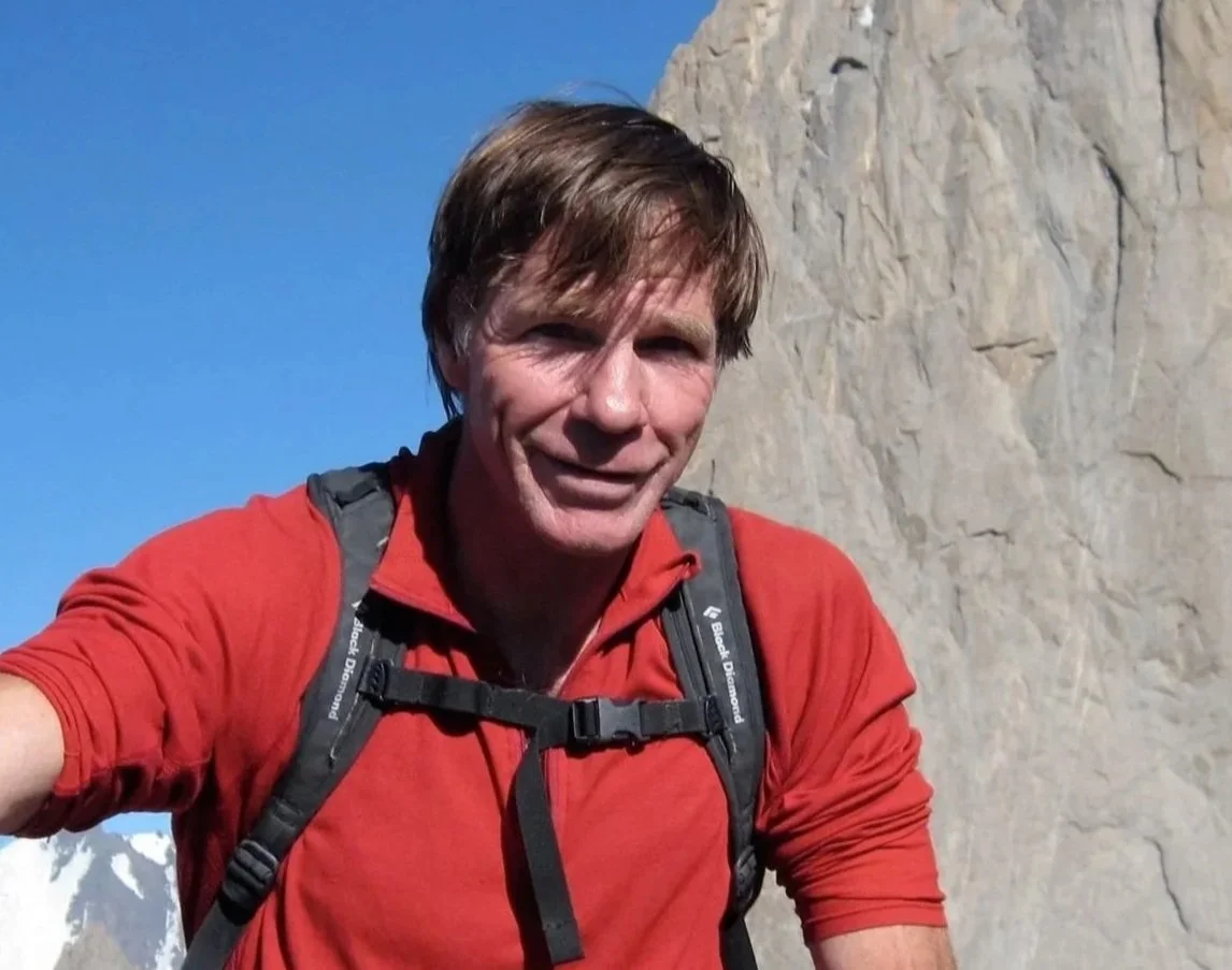 A man with brown hair wearing a red outdoor jacket and black backpack, smiling, in front of a mountain and clear blue sky.