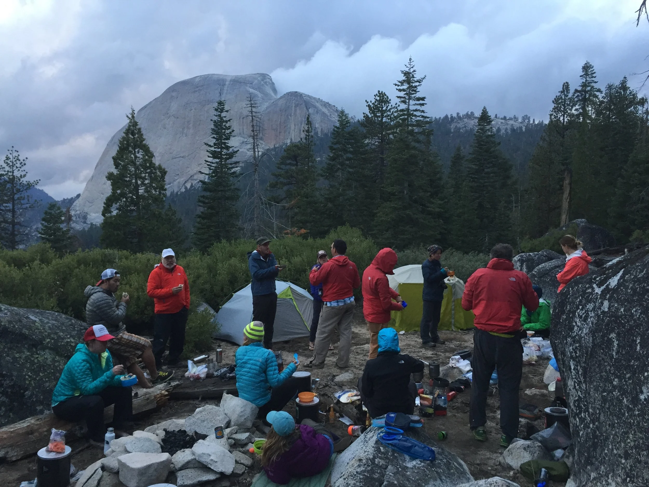 A group of people camping outdoors among trees and rocks with mountains in the background, some are sitting and some are standing, wearing outdoor clothing.