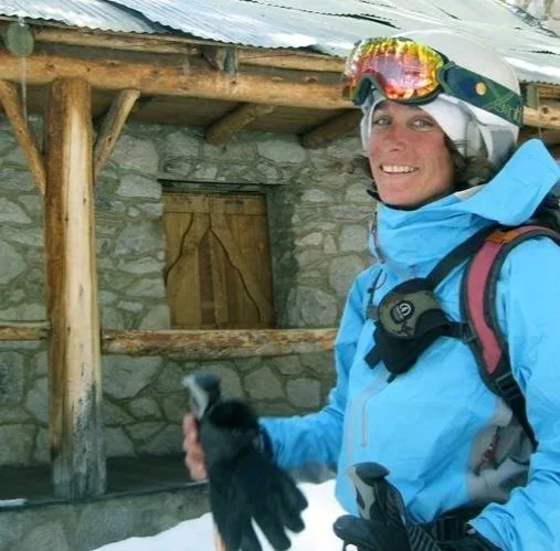 A woman in ski gear holding a penguin near a stone building with a wooden roof.
