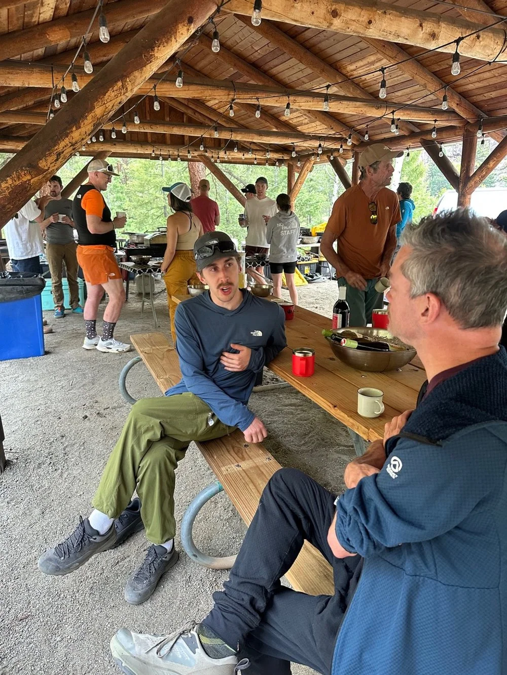 People gathered at an outdoor picnic shelter with a wooden roof, string lights, and a picnic table