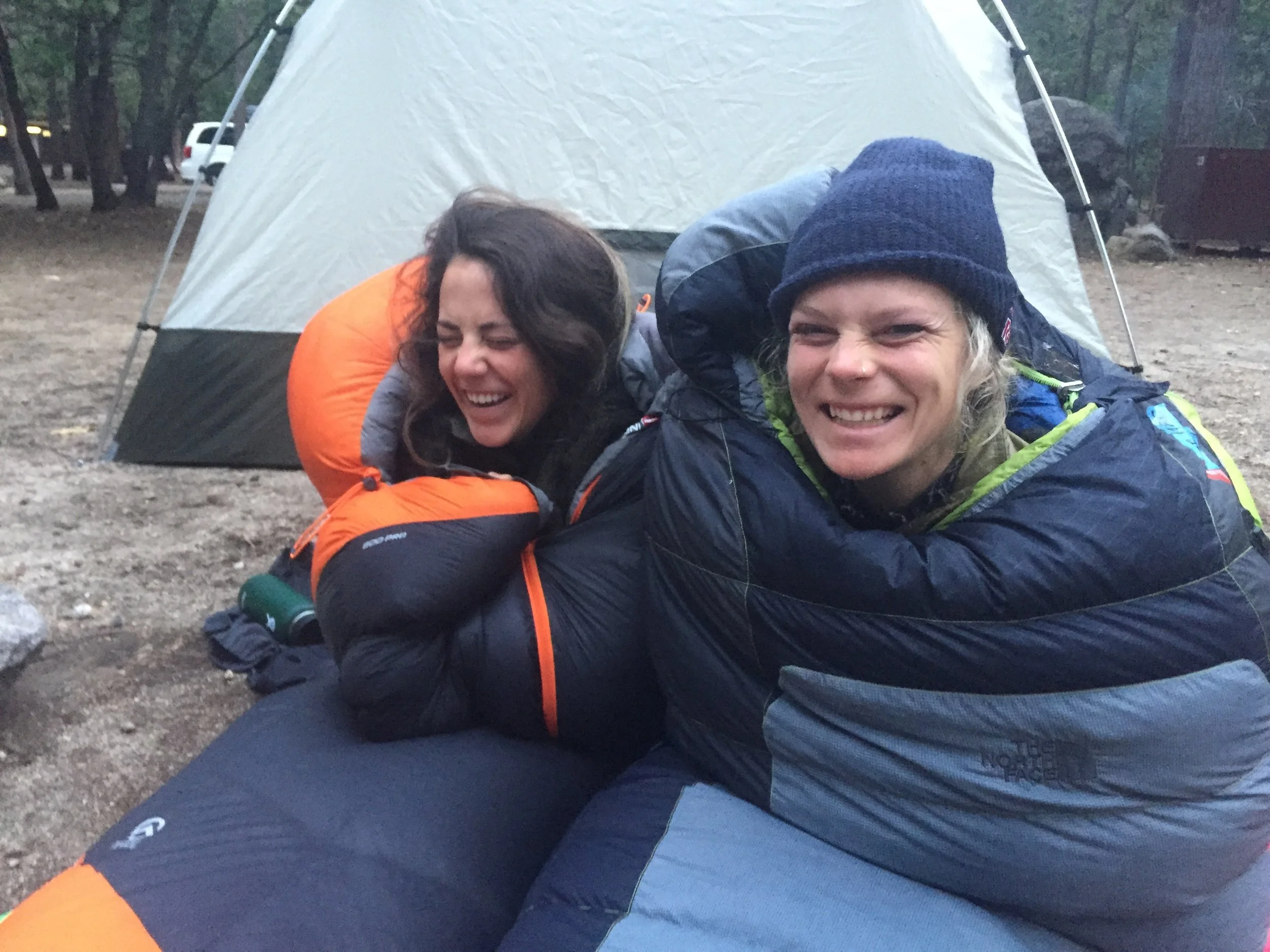 Two women in sleeping bags laughing and smiling outdoors near a tent in a wooded campsite.