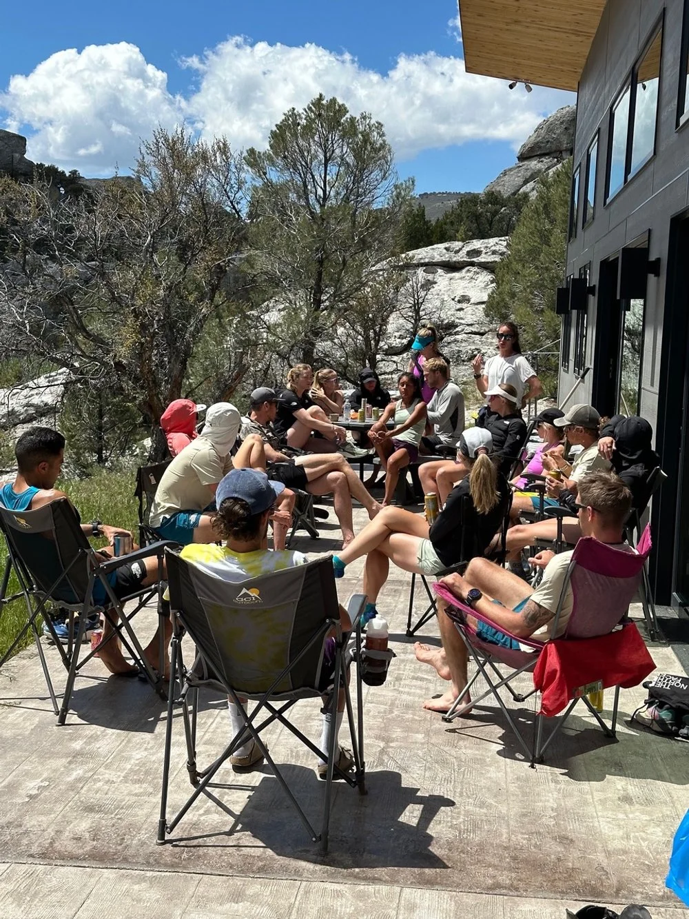 Group of people sitting on chairs outdoors on a patio, surrounded by trees and rocks with a partly cloudy sky overhead.