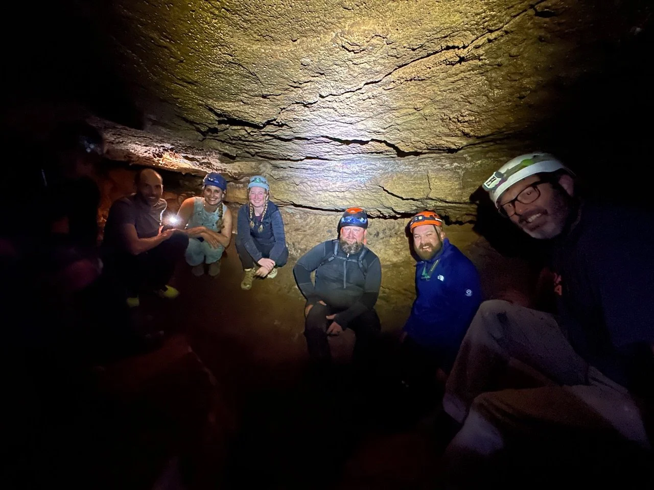 A group of six people wearing helmets and outdoor gear inside a dark cave, some with headlamps