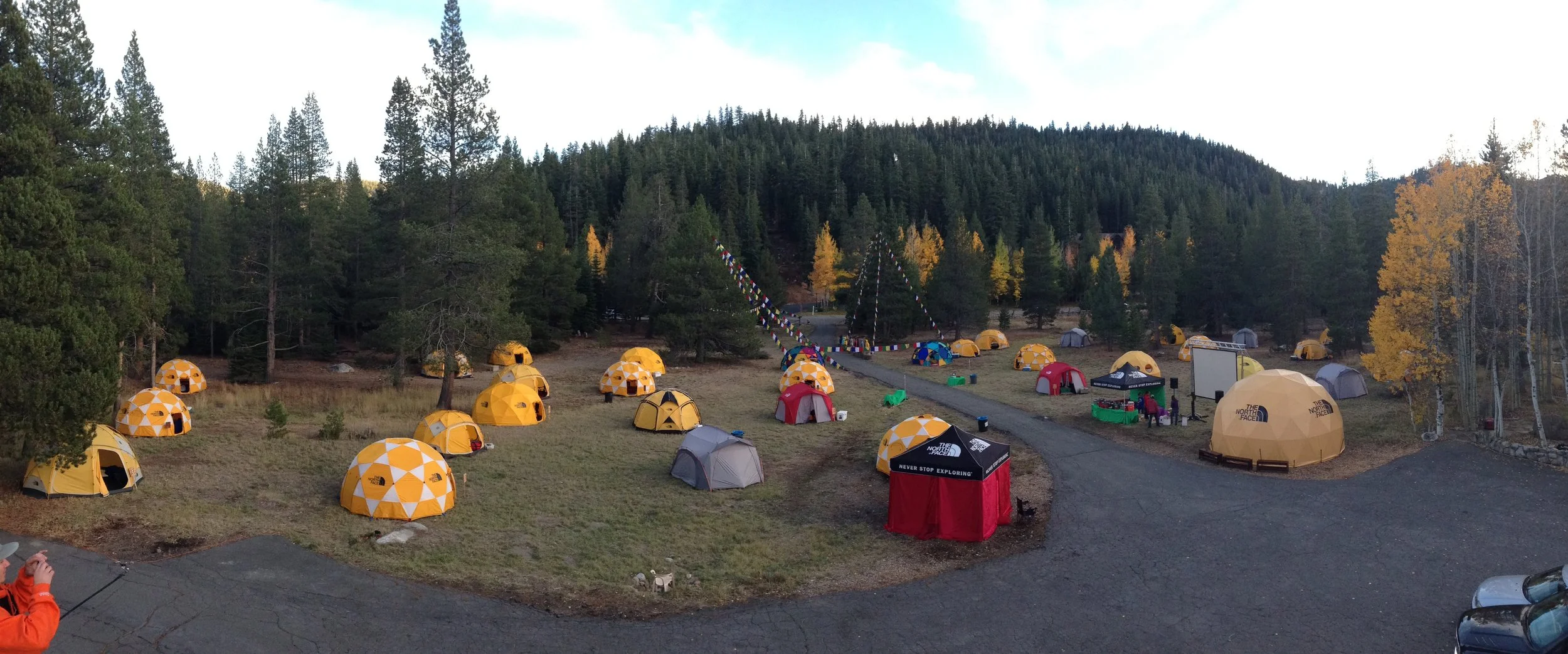 Campground with numerous yellow, grey, red, and blue tents set up in a forested area with tall pine and aspen trees, some with fall foliage, and a mountain in the background, decorated with colorful flags.