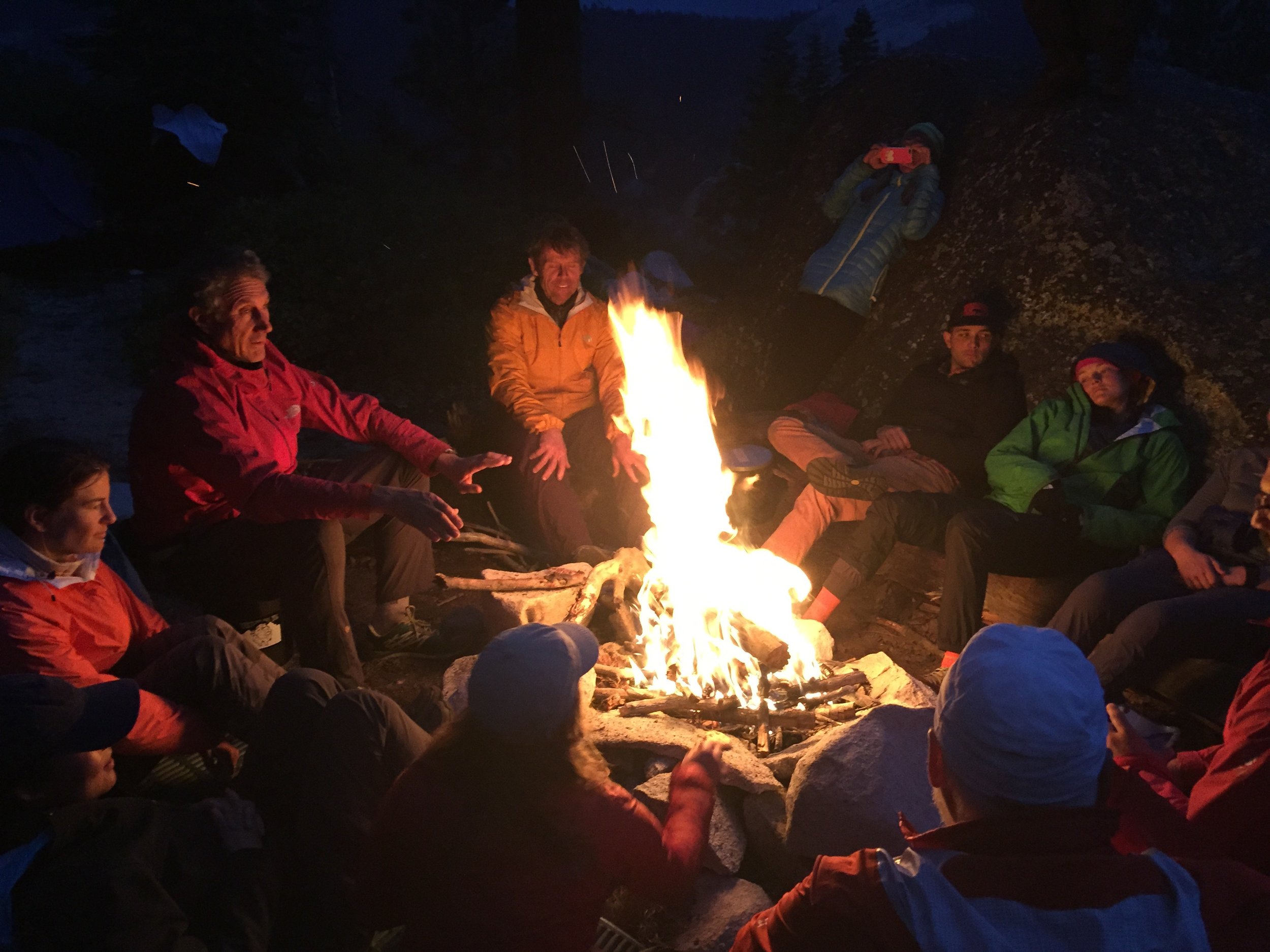 Group of people sitting around a campfire at night outdoors, wearing jackets and warm clothing, with some taking photos.