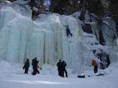 People ice climbing on a frozen waterfall in a snowy landscape.