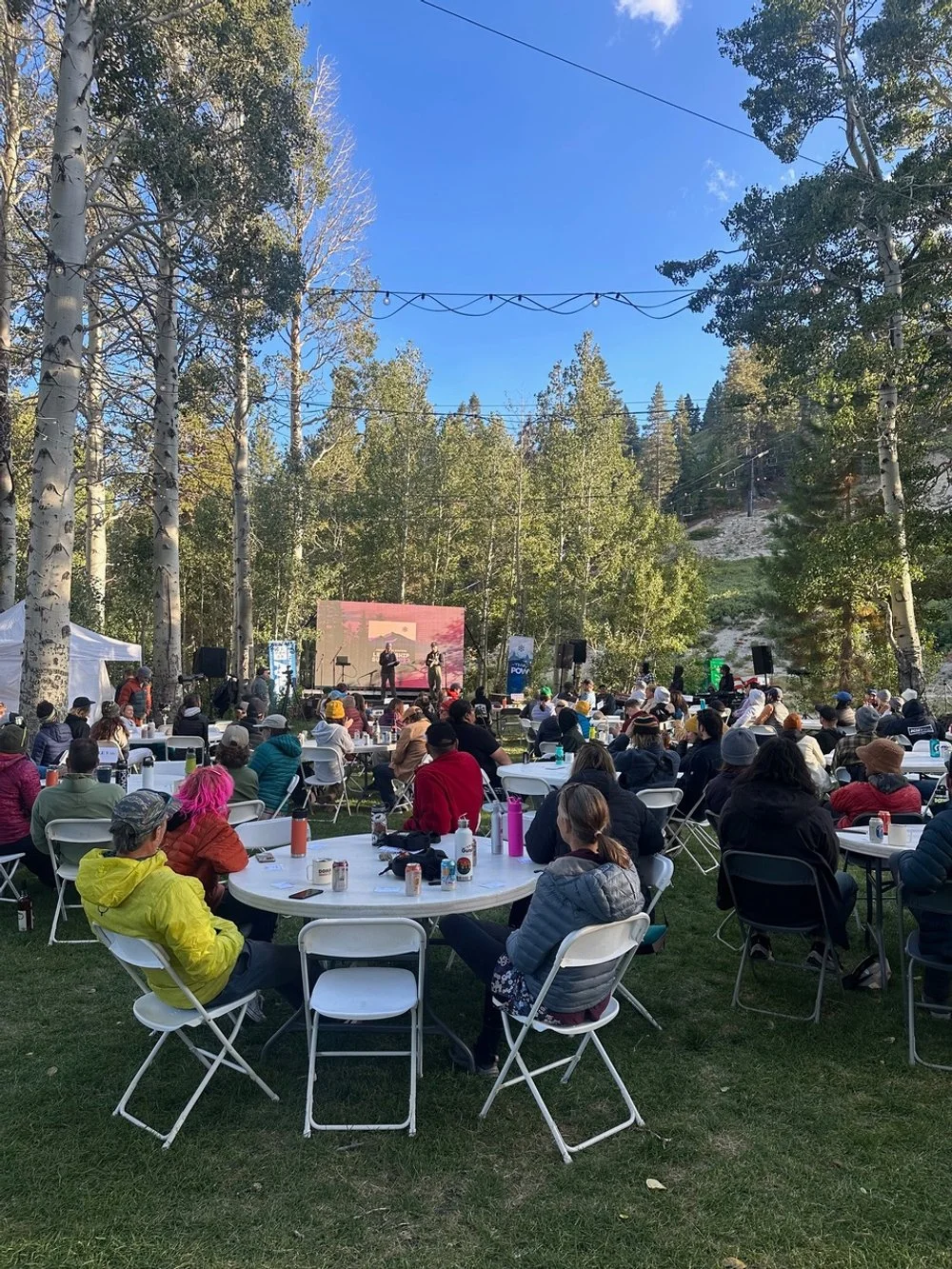 People attending an outdoor event in a forested area, sitting at round tables, listening to performers on a stage with a digital backdrop. 