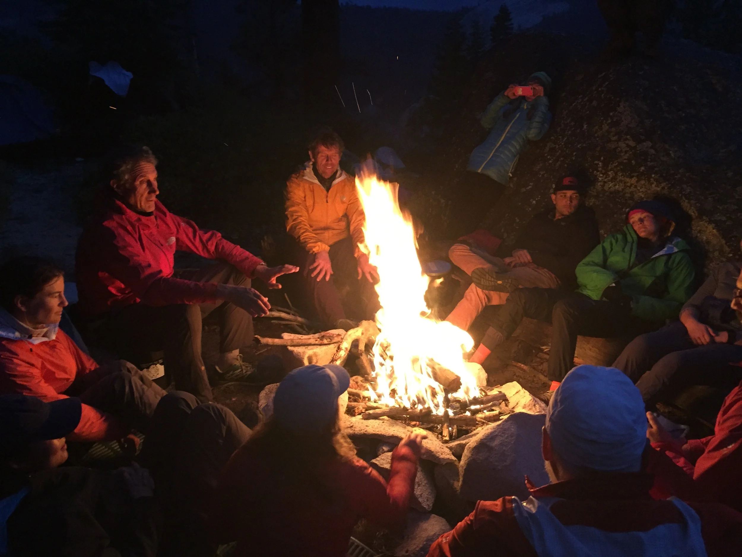 A group of people gathered around a campfire at night in an outdoor setting on a camping activity