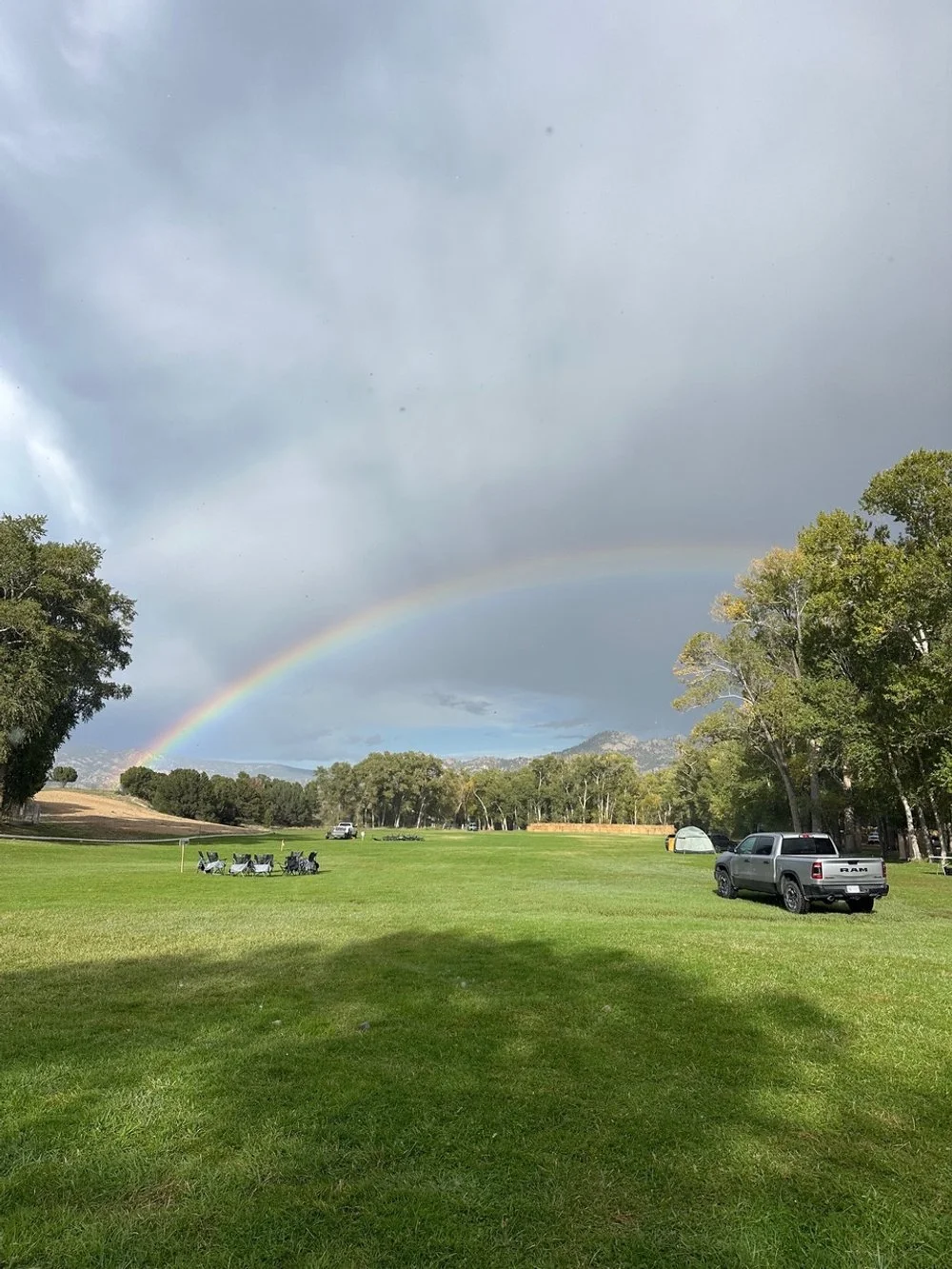 A park field with trees, parked cars, a tent, and a rainbow in a cloudy sky.