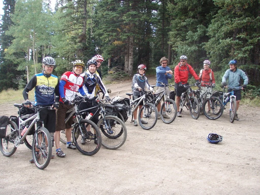 Group of nine people with bicycles on a dirt trail in a forested area, wearing helmets and cycling gear, some smiling and others with arms crossed.