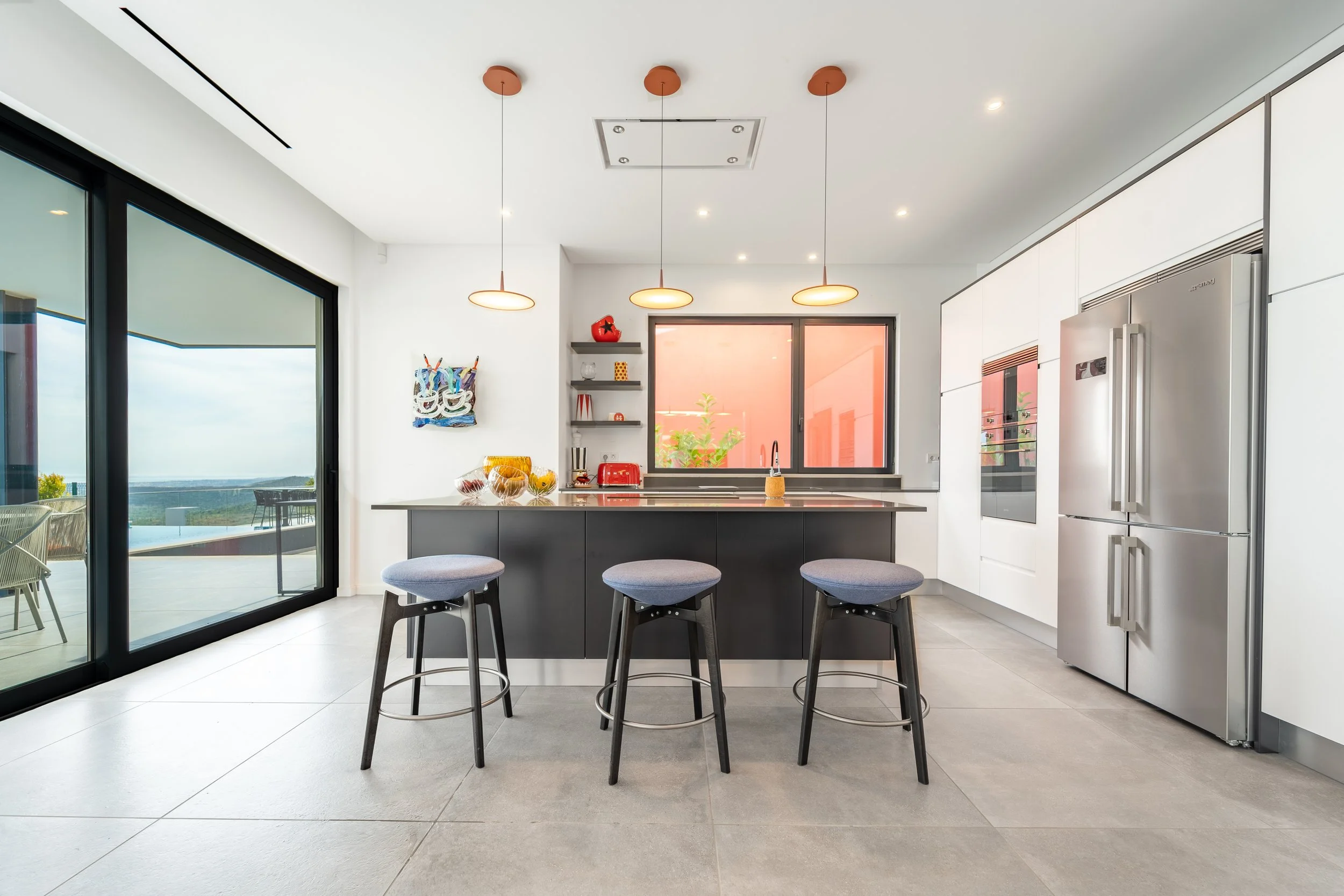 Cozinha moderna com ilha central, três banquetas de bar, geladeira de aço inox, armários brancos, janela com vista para o exterior, prateleiras decorativas, eletrodomésticos vermelhos, iluminação pendente, piso de cerâmica.