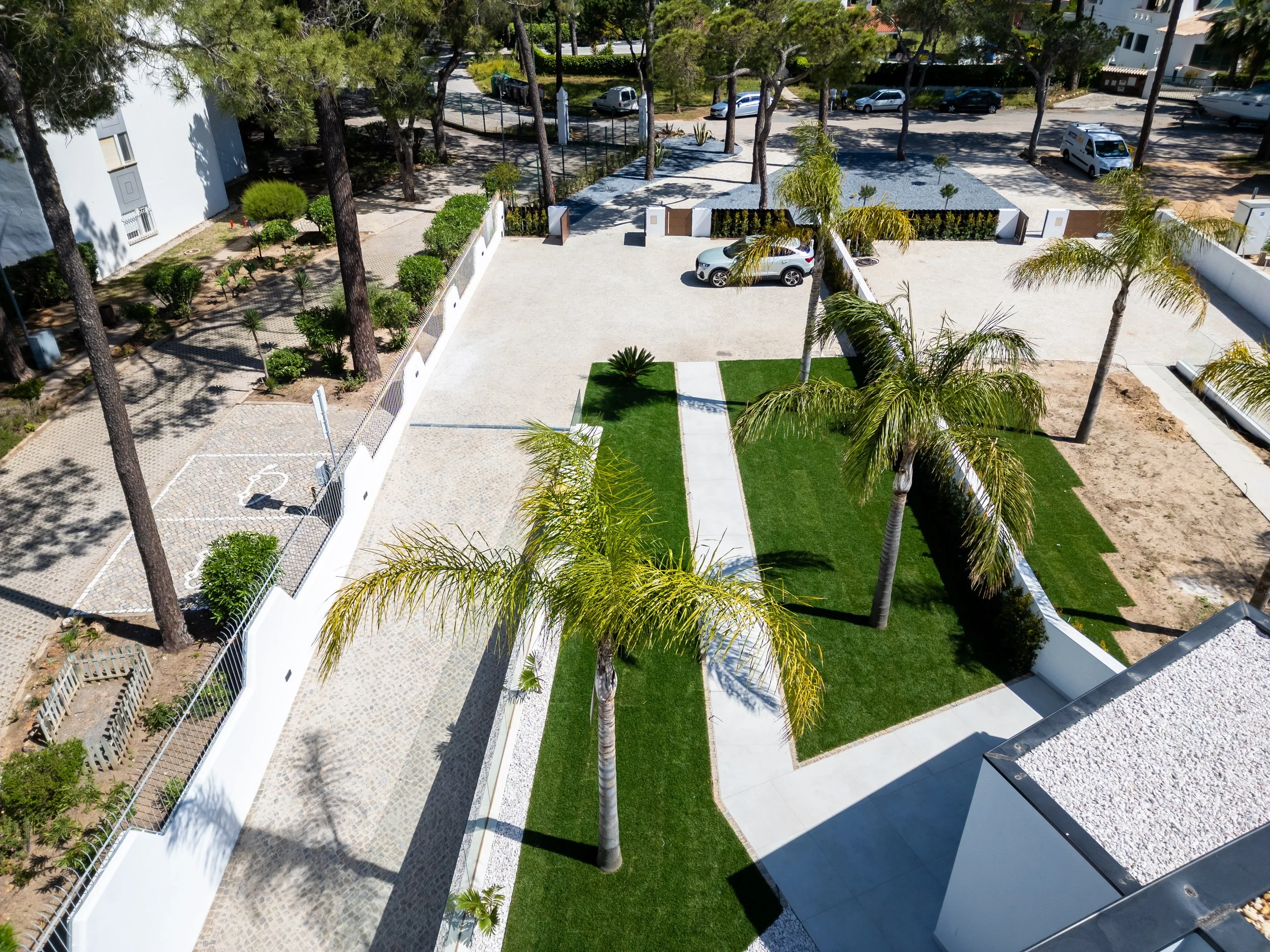 Vista aérea de uma área residencial com jardim bem cuidado, árvores palmeiras, calçadas de concreto e algumas áreas de estacionamento com carros. Há uma área gramada e uma calçada central que conecta diferentes partes do espaço.