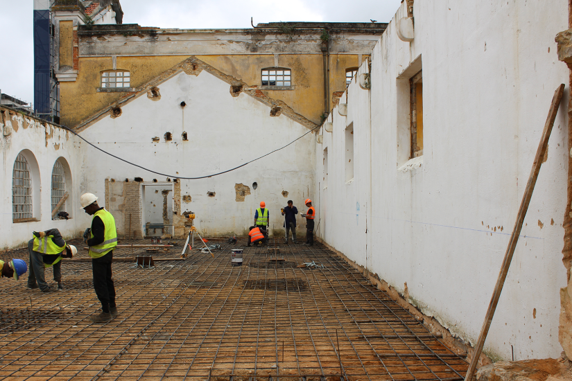 Obra de construção com trabalhadores colocando reforço de aço no chão, cercada por paredes antigas e uma estrutura de edifício ao fundo.
