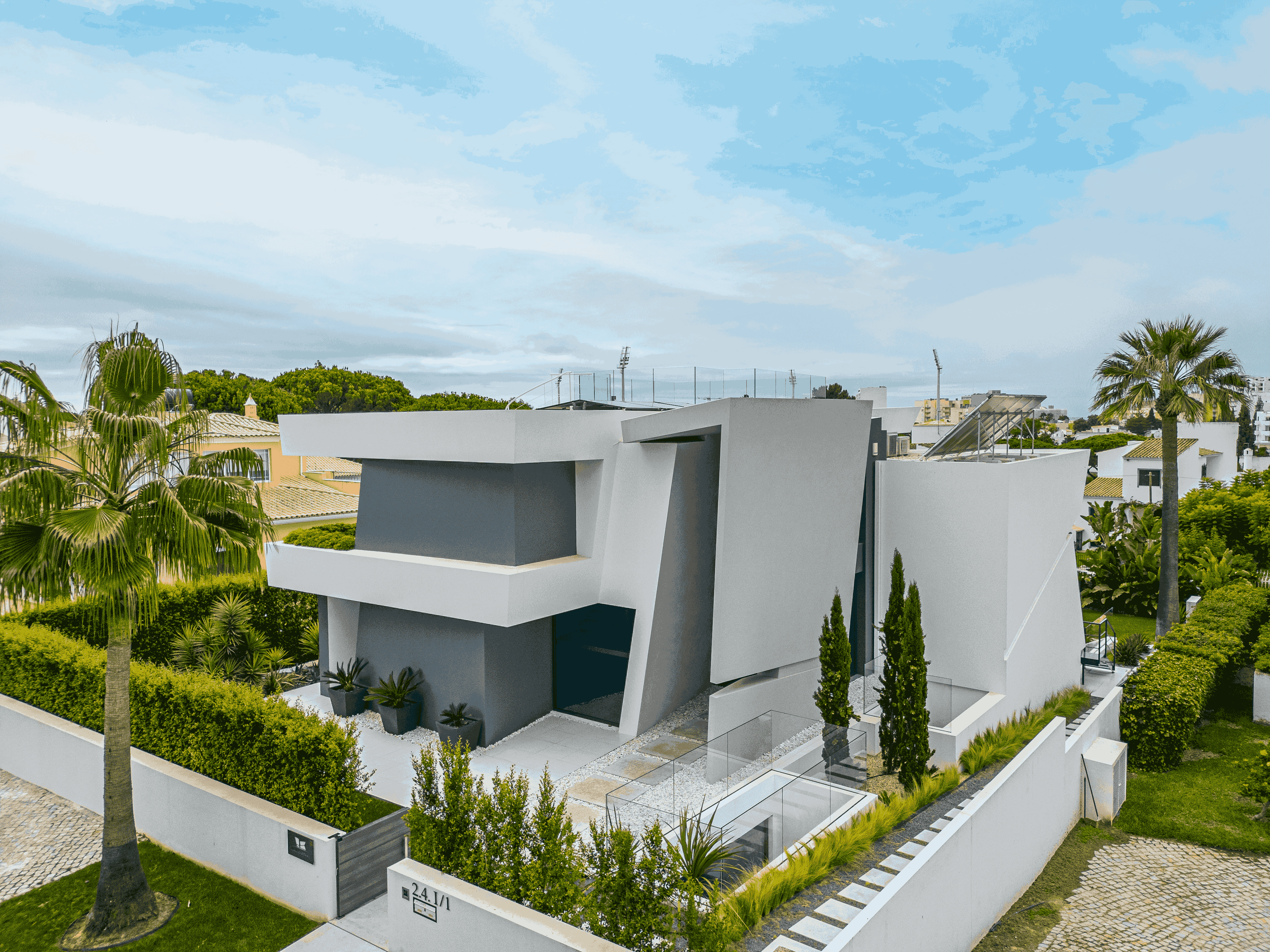 Casa moderna de cor branca com detalhes cinza, rodeada por vegetação e árvores, com muro branco, portão e calçada de pedra, sob céu parcialmente nublado.