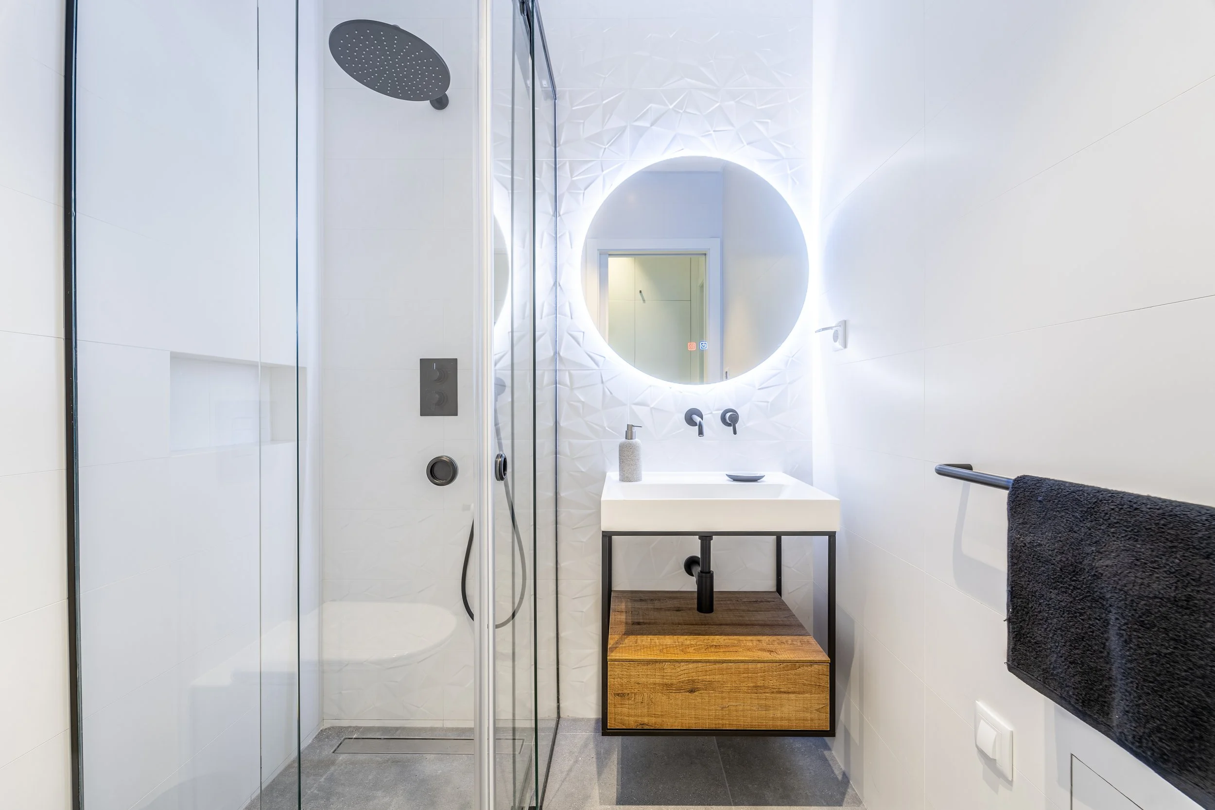 Banheiro com pia, espelho com iluminação de LED, chuveiro de teto, parede branca texturizada, toalha preta, paredes brancas e piso cinza.