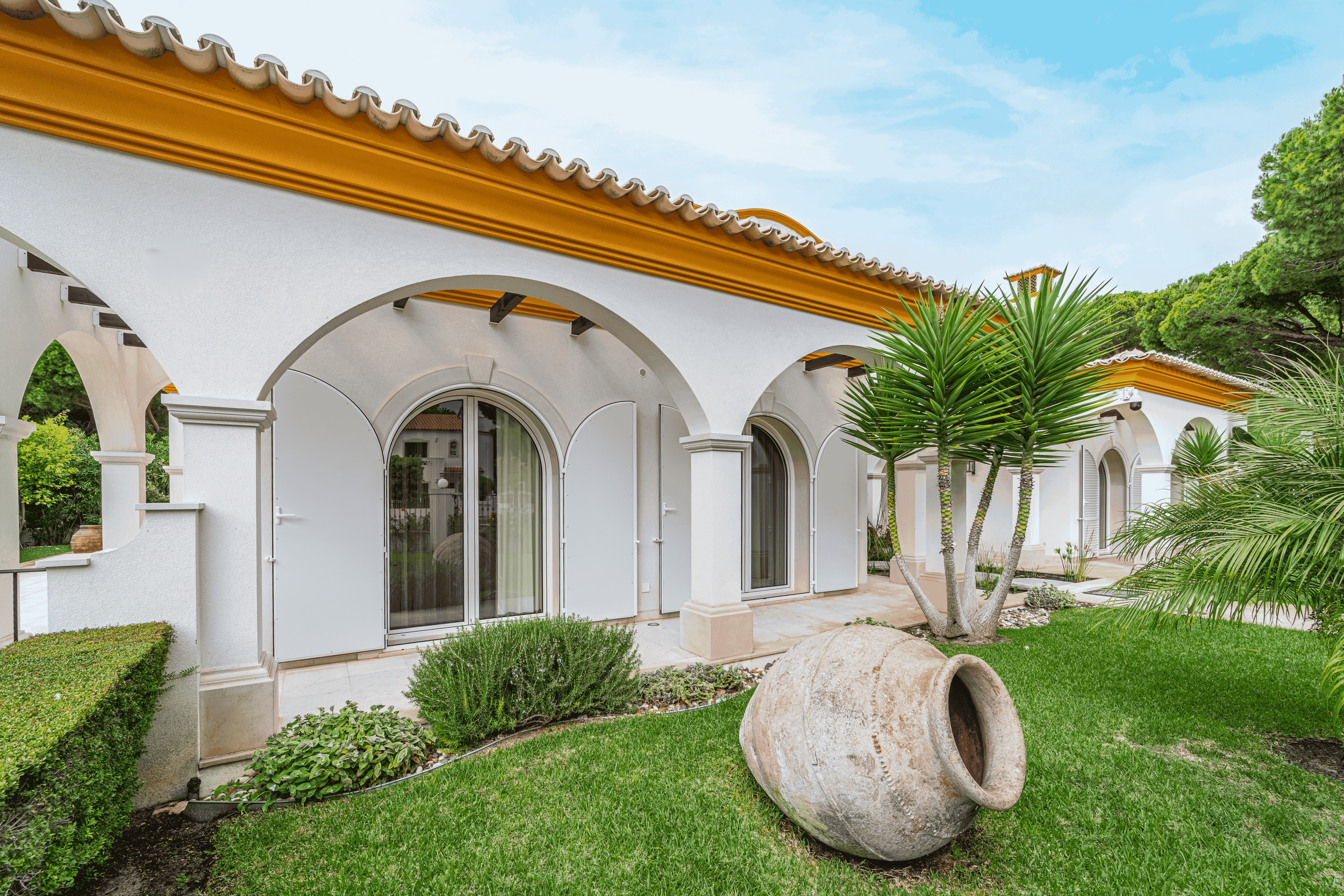 Casa com jardim, árvores e vaso de barro no gramado, sob céu claro.