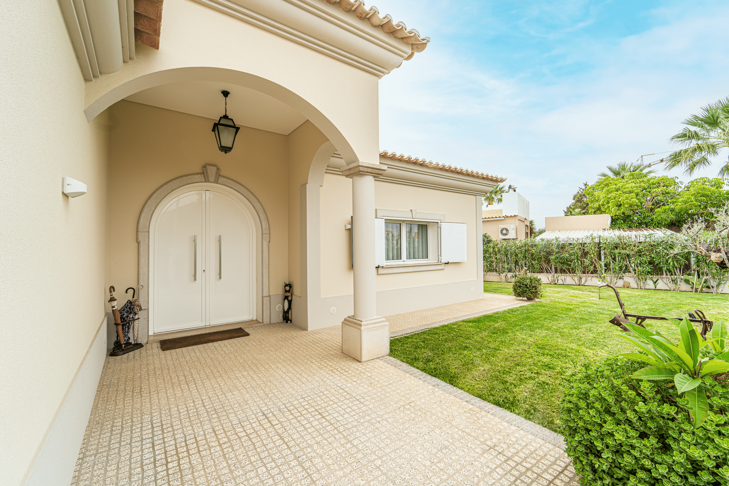 Fachada de uma casa com varanda, porta de entrada branca com arco, jardim bem cuidado, árvores e arbustos, céu azul com nuvens, piso de pedra e cadeira de jardim no quintal.