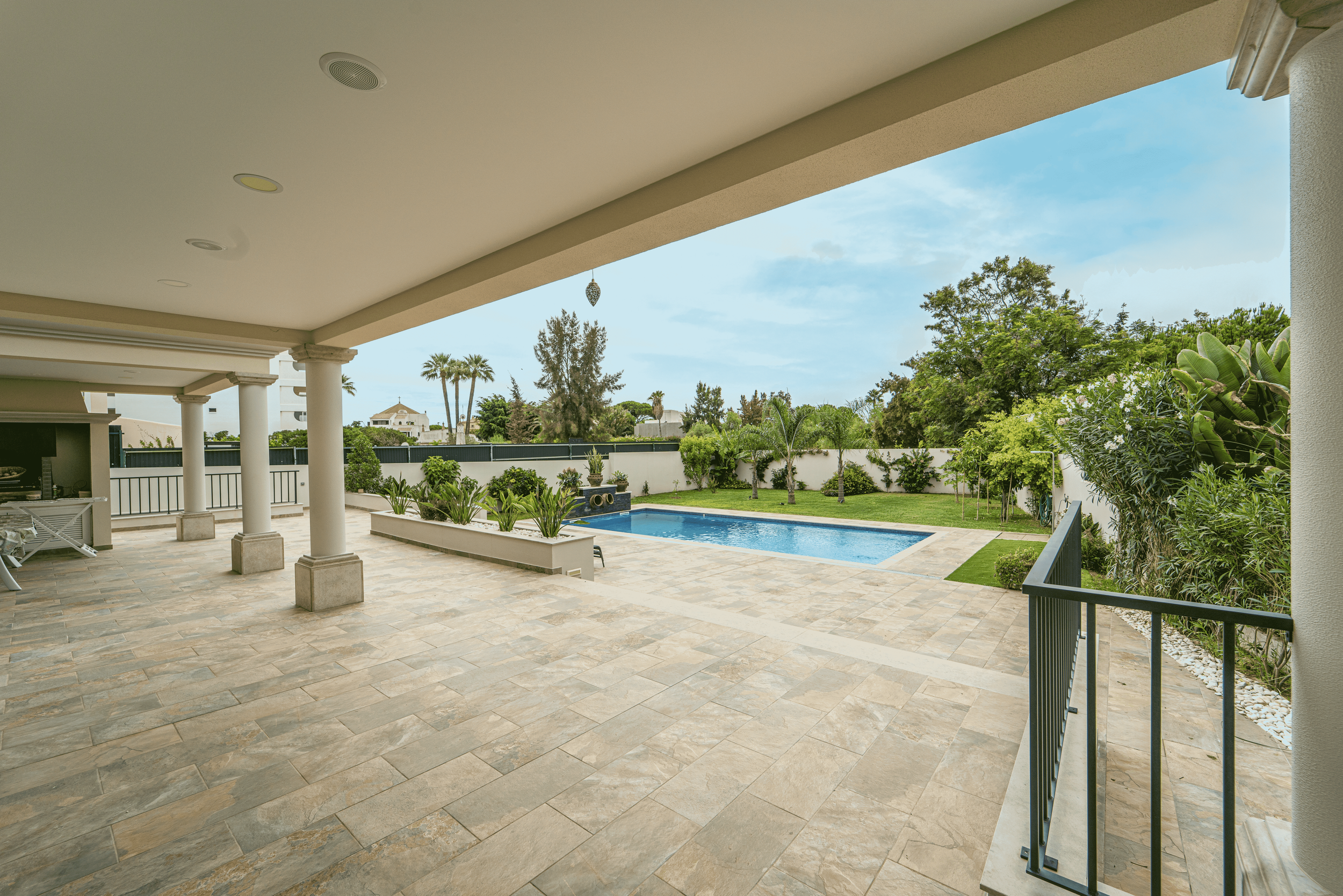 Varanda de casa com piso de pedra, vista para piscina de água azul, jardim com árvores e plantas ao redor, céu parcialmente nublado.