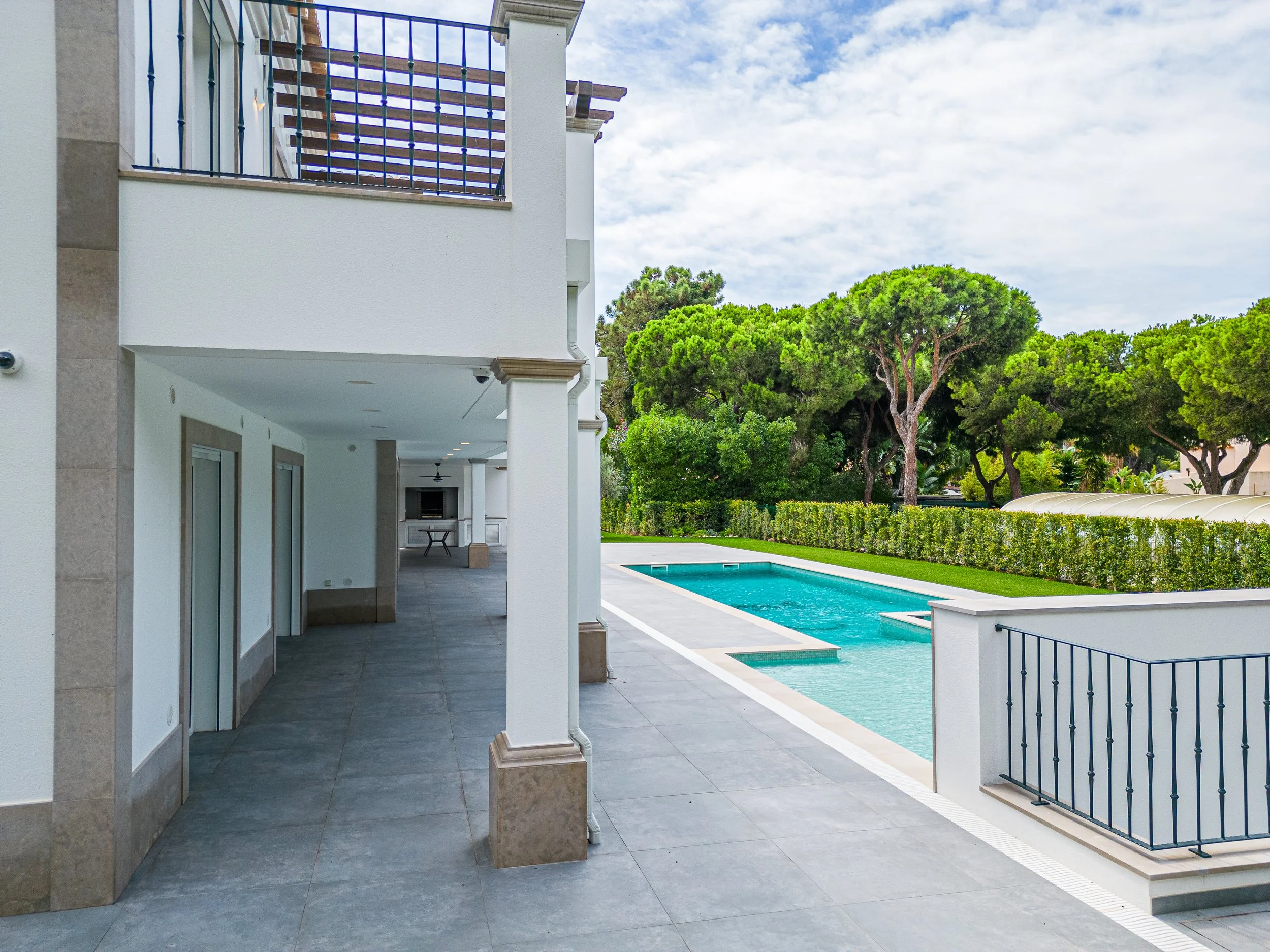 Área de piscina em um espaço exterior de uma residência moderna com árvores ao fundo, parede branca, piso de azulejos cinza e cerca de ferro ao lado da piscina.