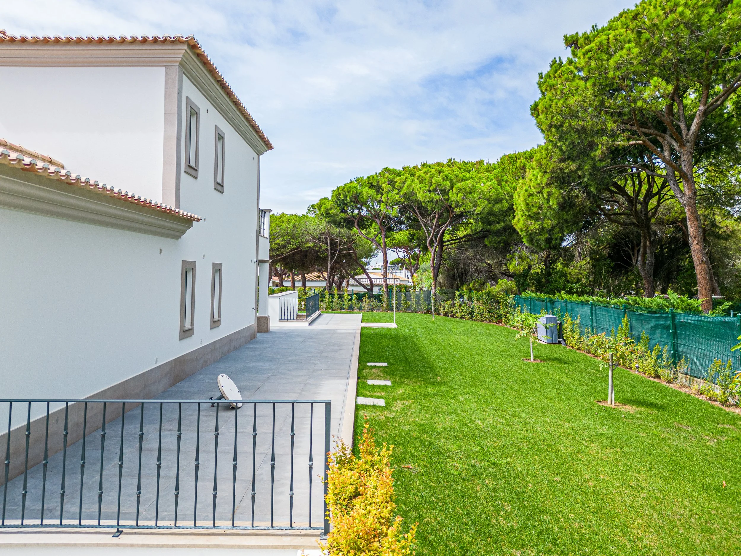 Jardim bem cuidado ao lado de uma casa branca com árvores verdes ao fundo e céu azul.