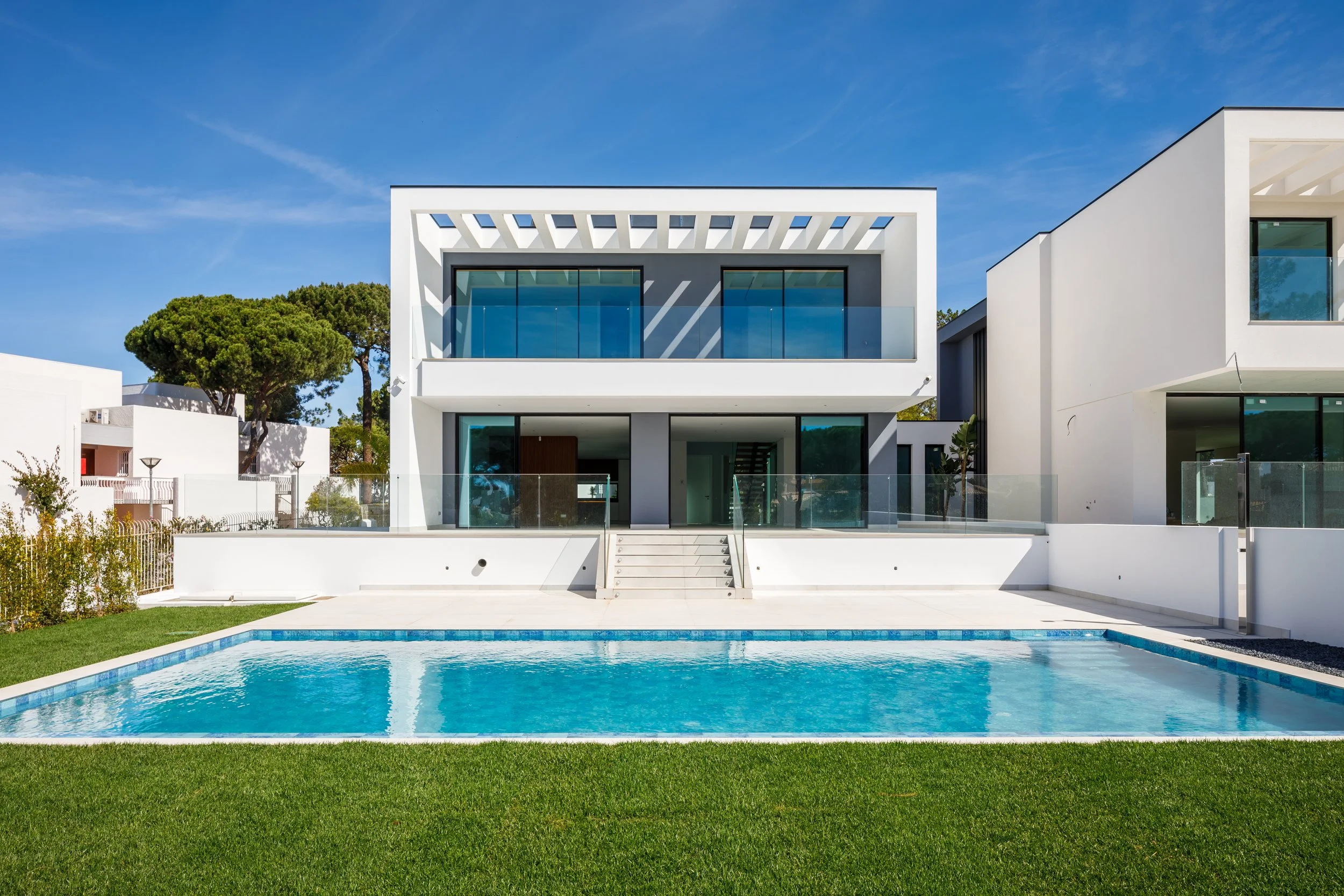 Casa moderna branca com janelas de vidro, piscina de prova azul e jardim verde sob céu azul.