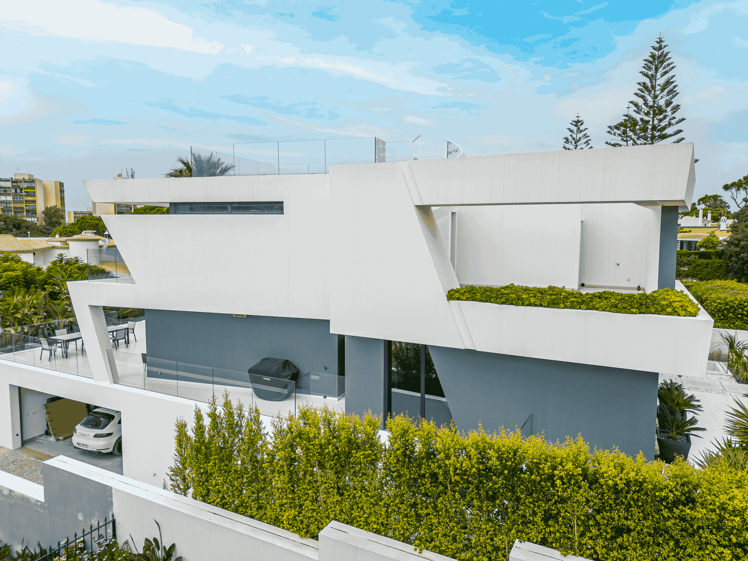 Prédio moderno branco com estrutura angular, varandas com vidro e área com jardim no topo, ao fundo céu azul com nuvens e árvores altas.